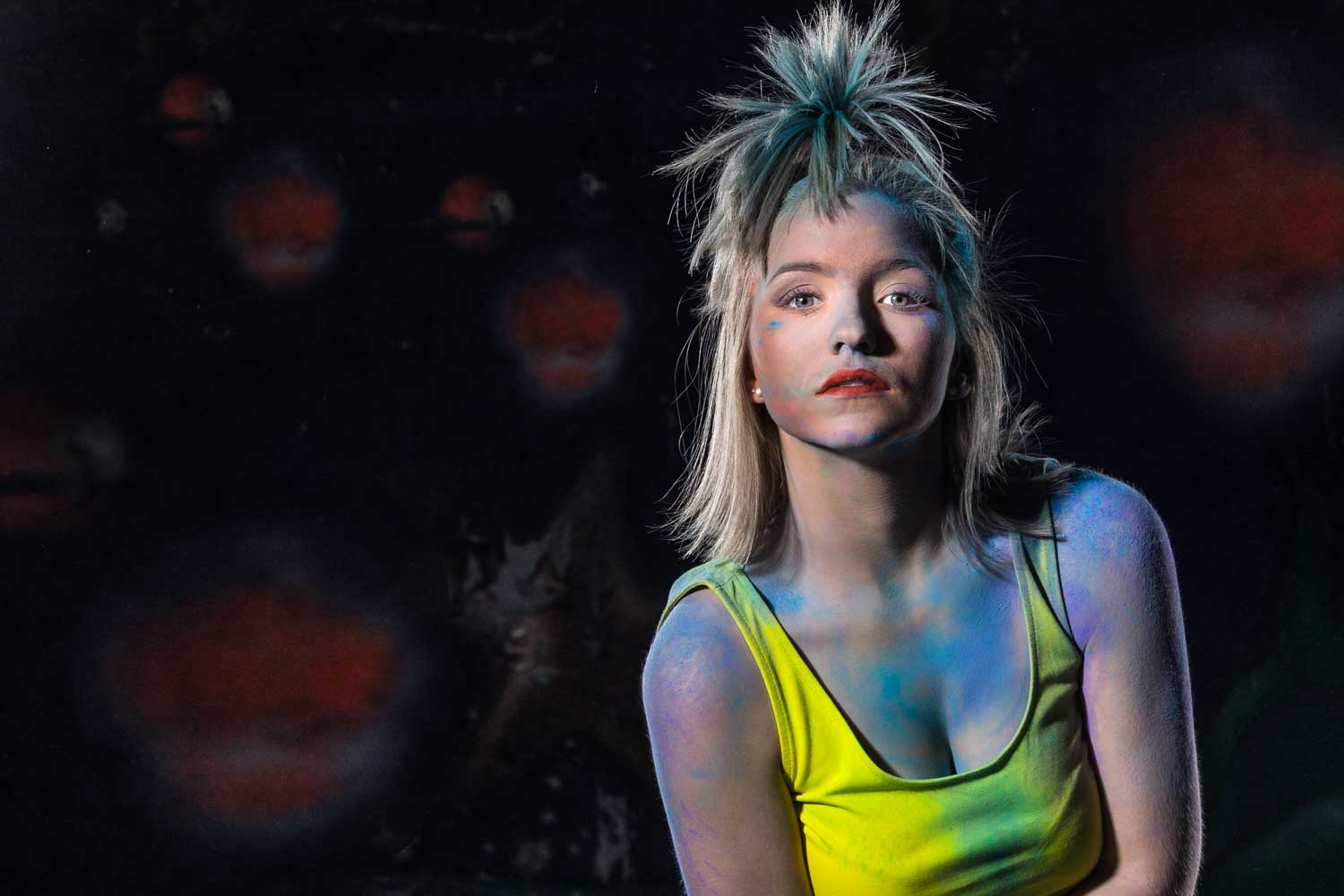 Woman with colorful paint on face and clothes, wearing a yellow top against a dark, abstract background.