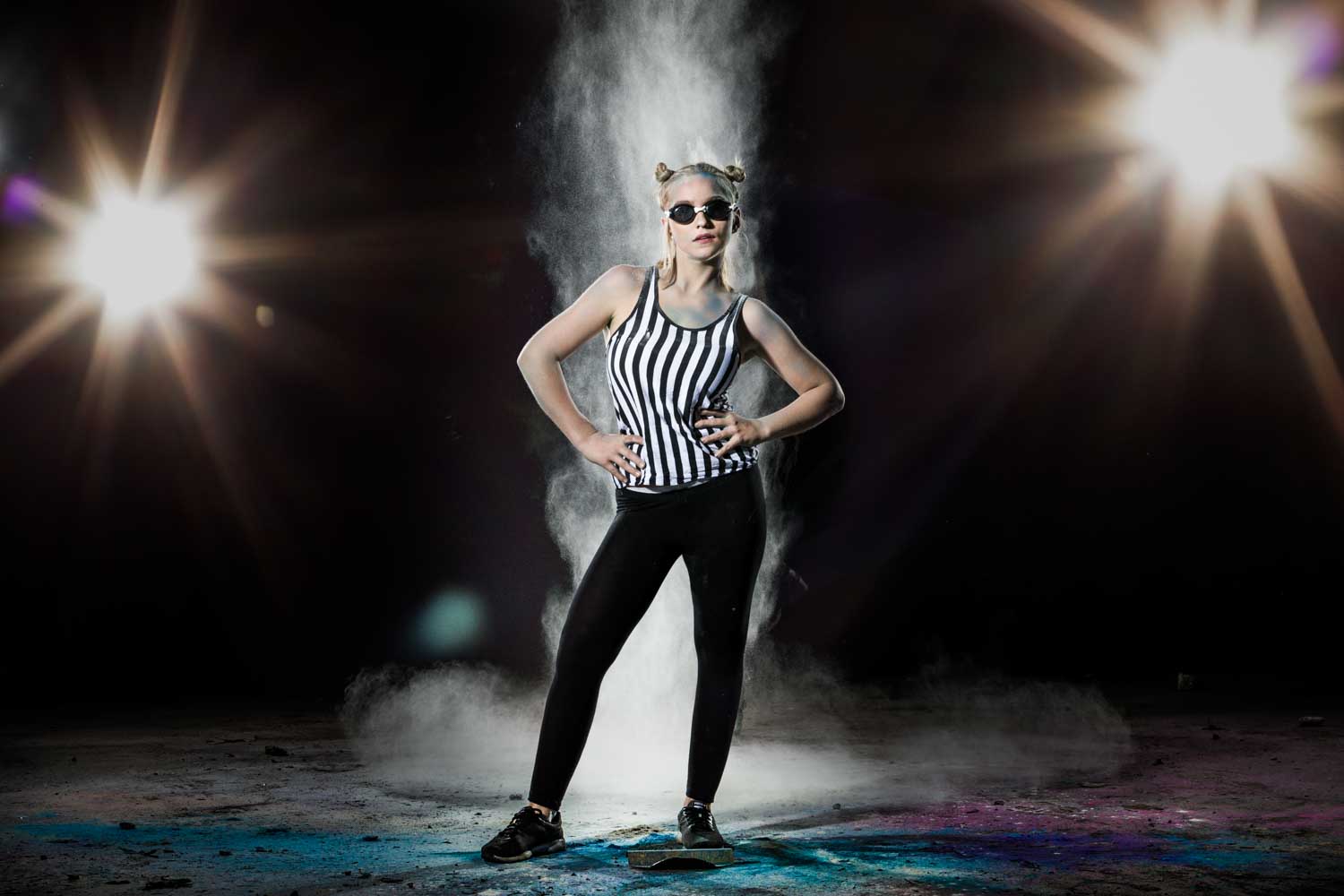 Stylish woman in stripes and shades poses confidently with dramatic lighting and dust effects in a dark setting.