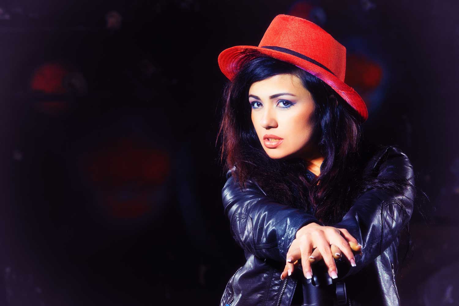 Woman in a black leather jacket and red hat poses confidently against a dark background.