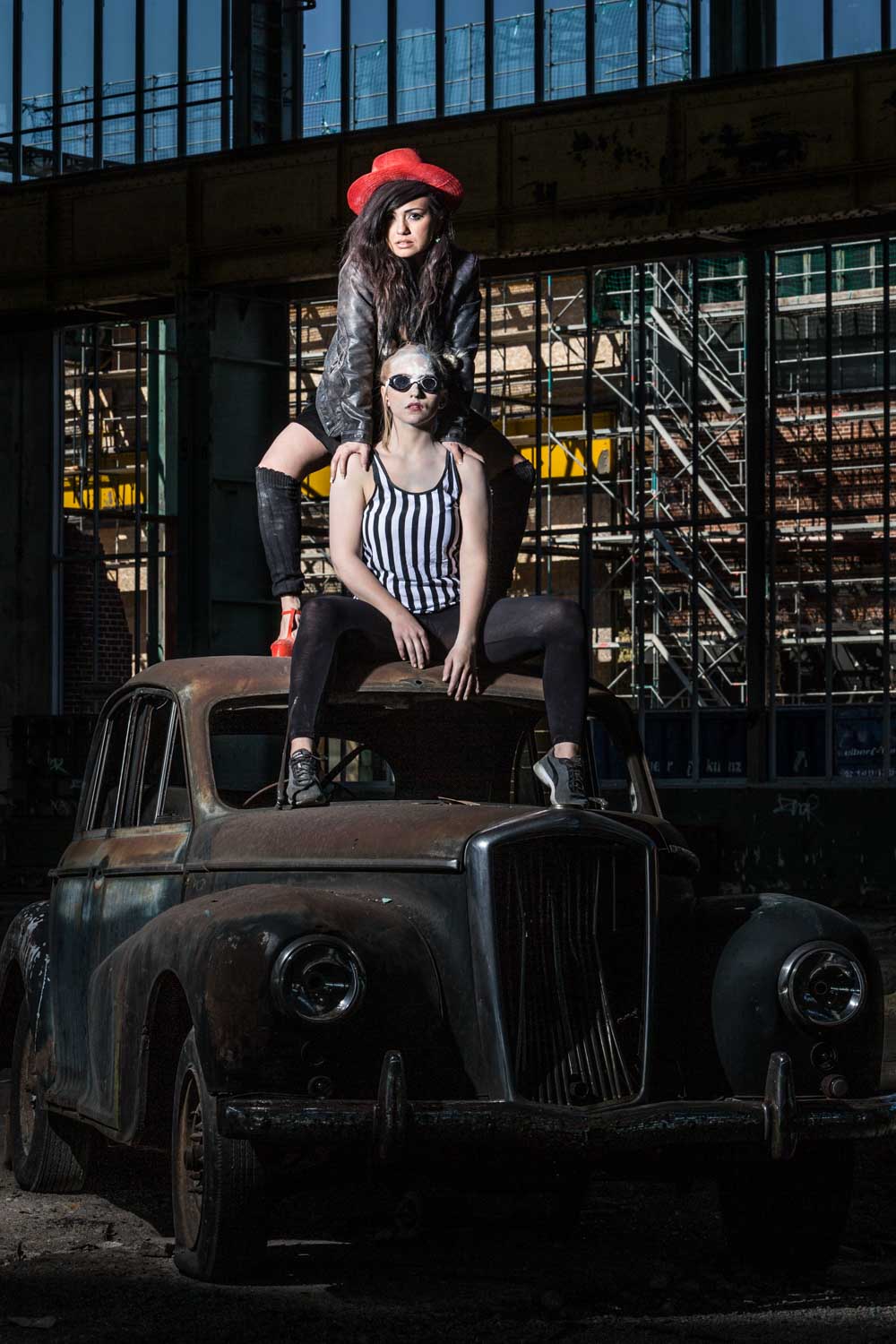 Two stylish women pose on a vintage car in an industrial setting, one wearing a red hat, the other in a striped top.