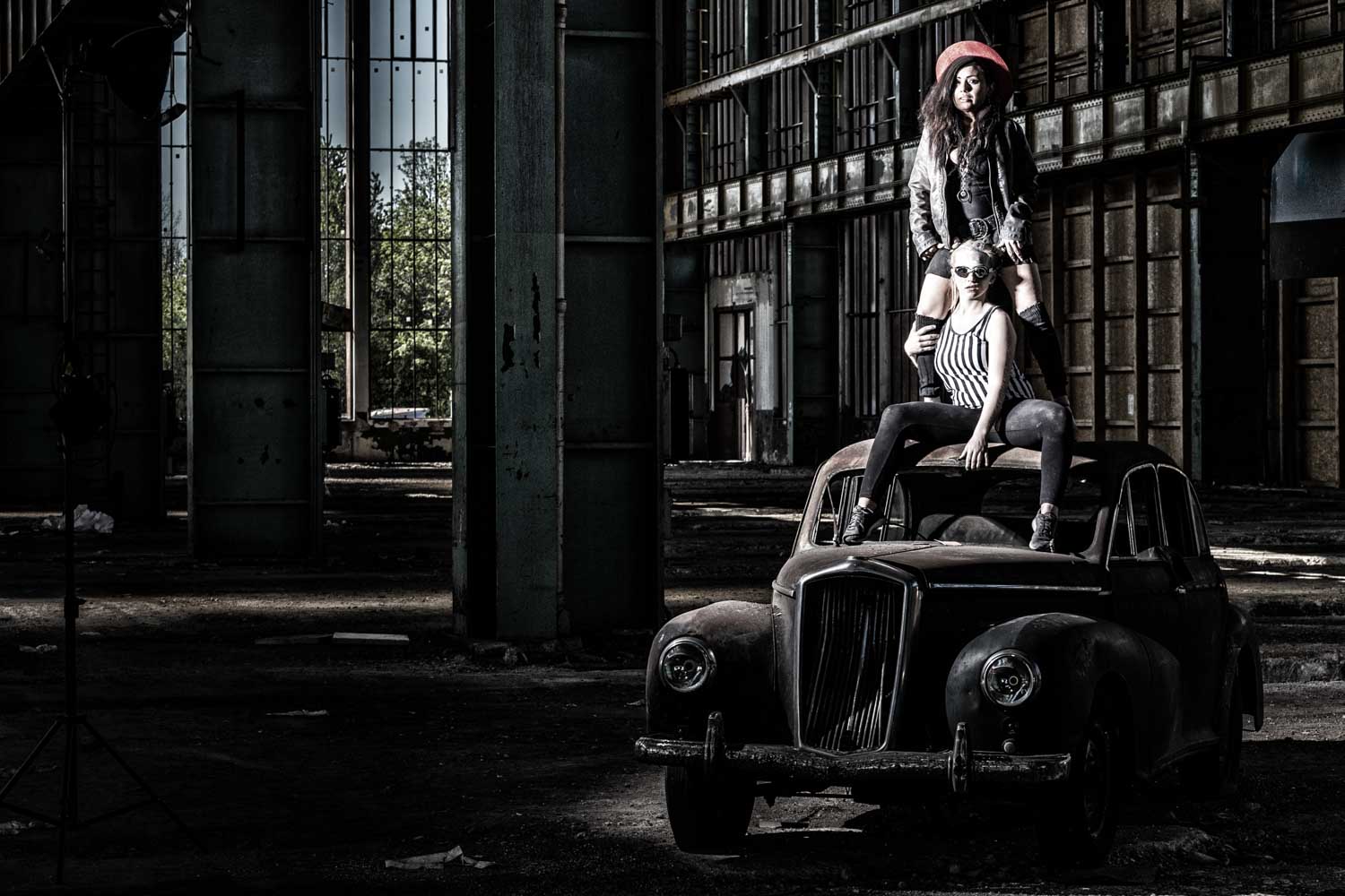 Two women posing on a vintage car in an industrial warehouse setting, highlighting fashion and urban vibes.