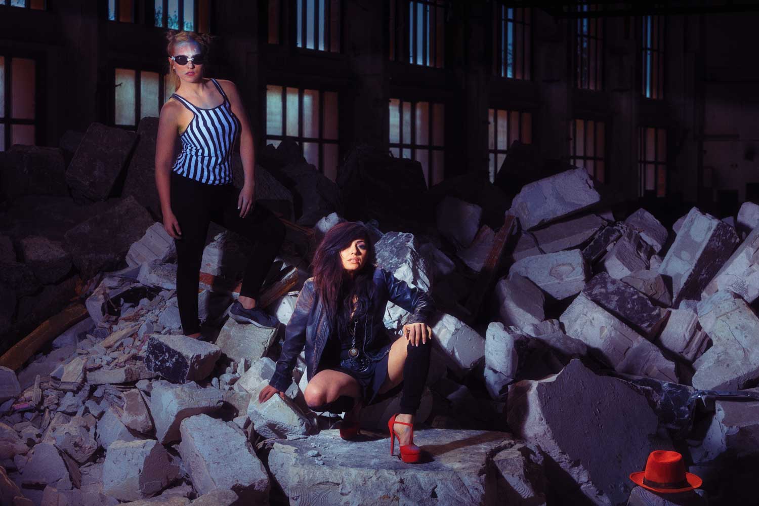 Two fashionable women pose on rubble in a dark warehouse, wearing a striped top and leather jacket with red high heels.