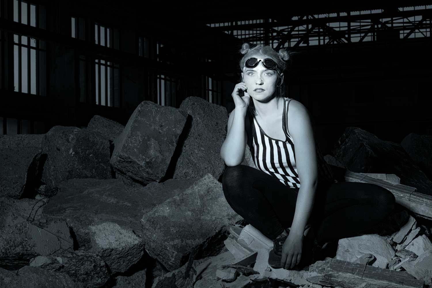 Woman in striped tank top and goggles sitting on rubble in dimly lit warehouse.