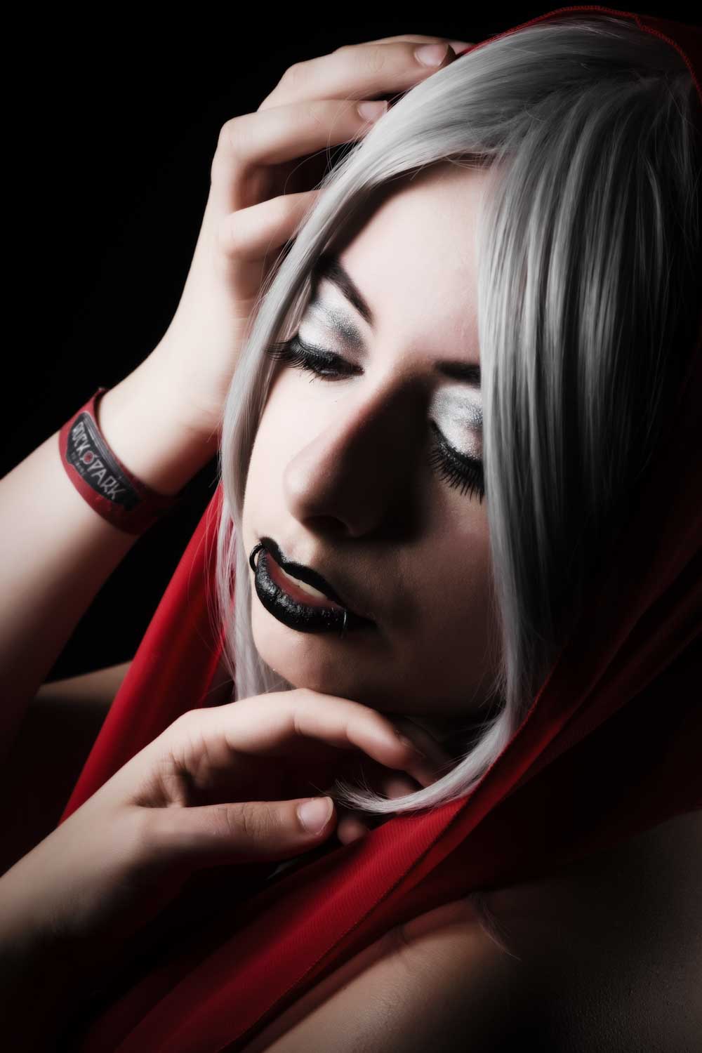 Person with silver hair, dramatic makeup, and red hood posing against a dark background, expressing gothic style.