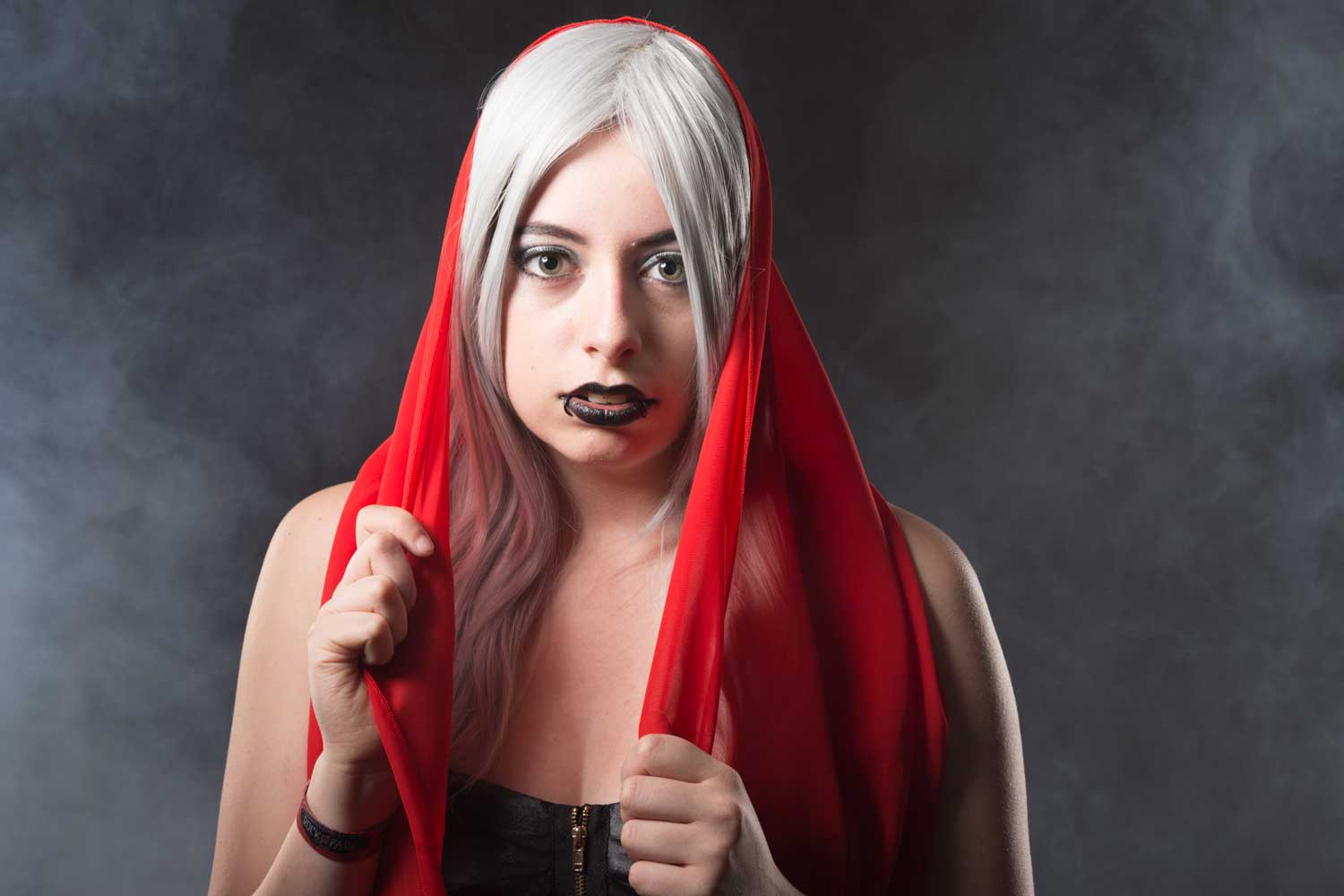 Woman with white hair and black lipstick wearing a red veil, in front of a smoky background.