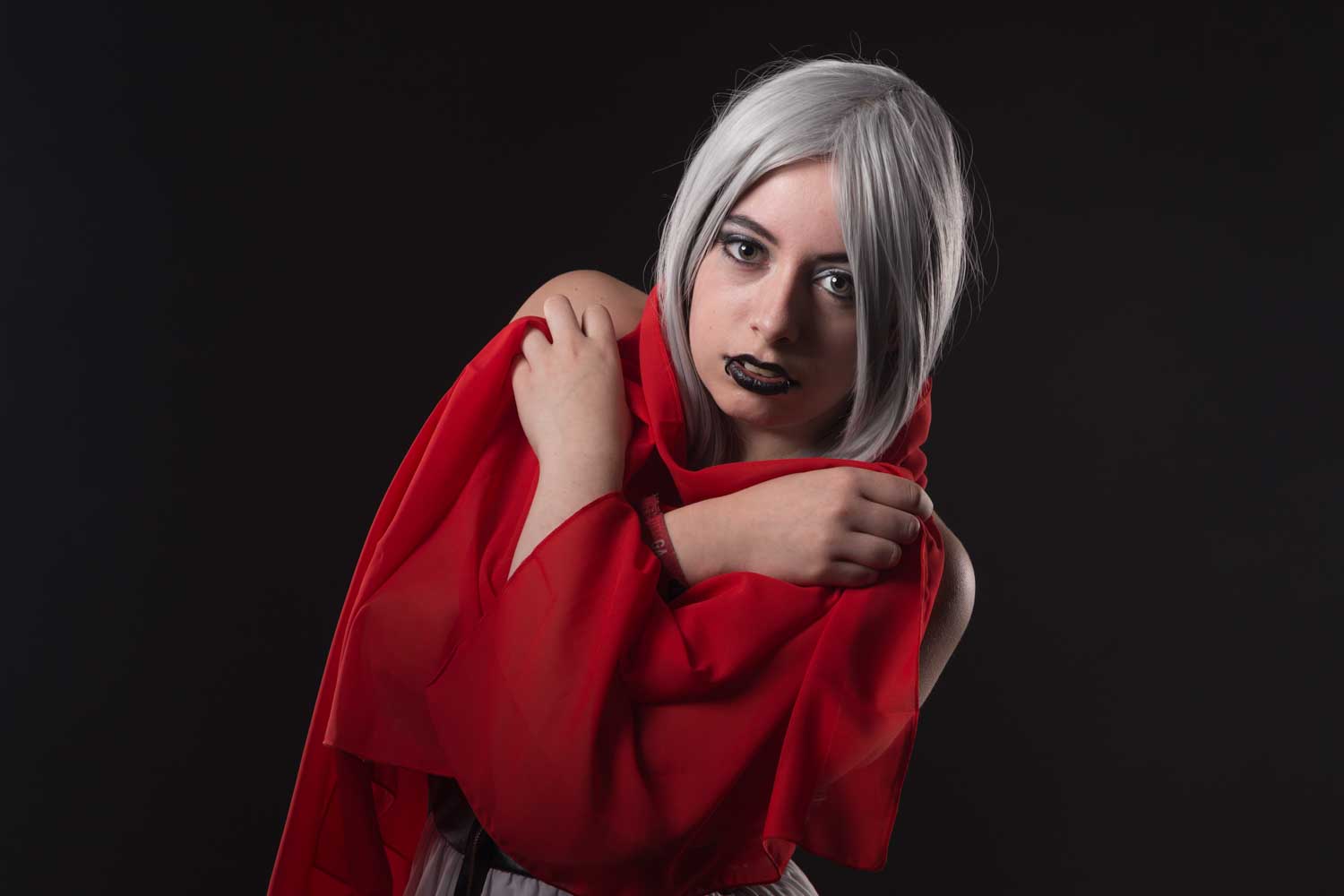 Young person with white hair draped in red fabric, wearing black lipstick, against a dark background.