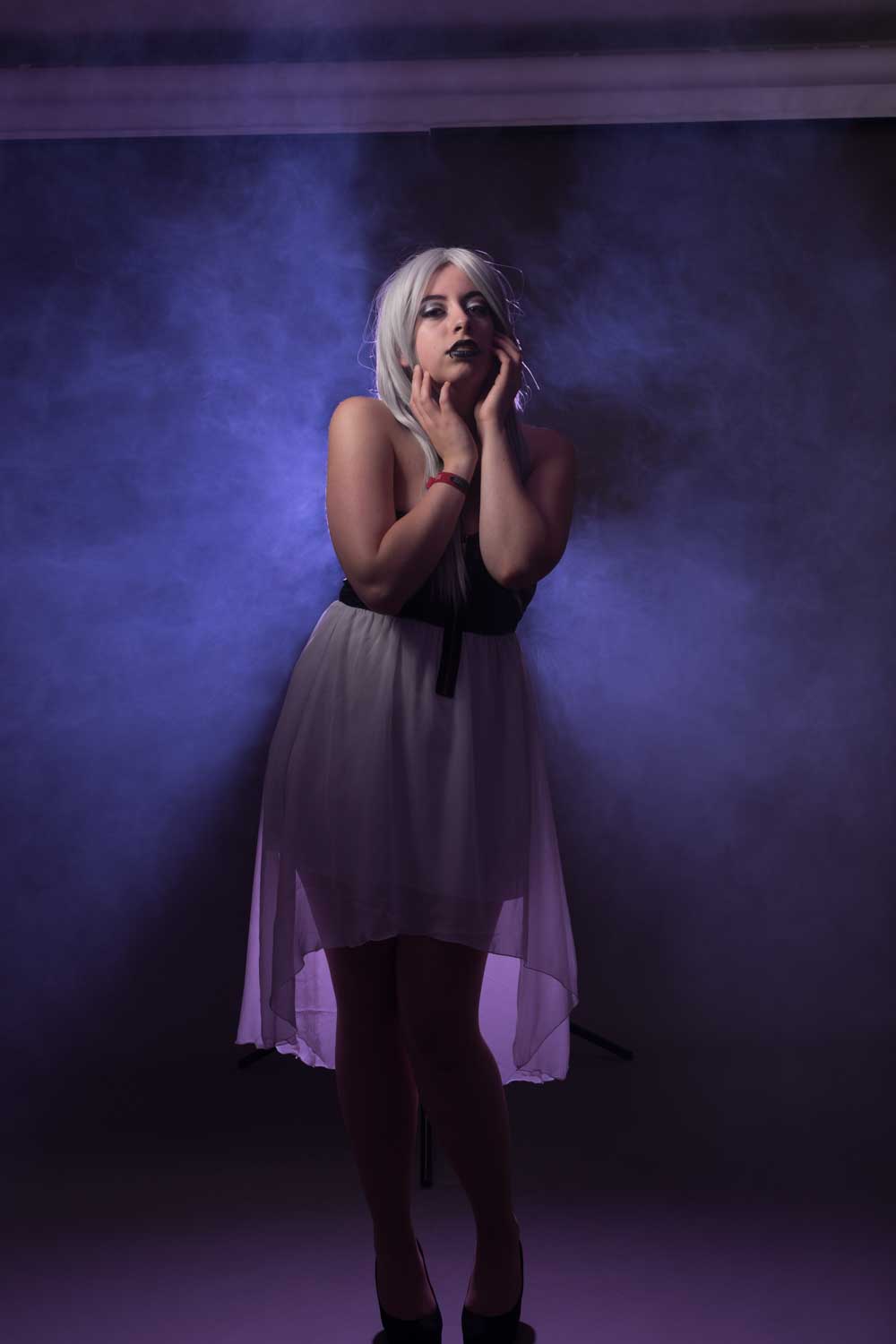 Woman with white wig in sheer dress posing amid purple foggy backdrop.