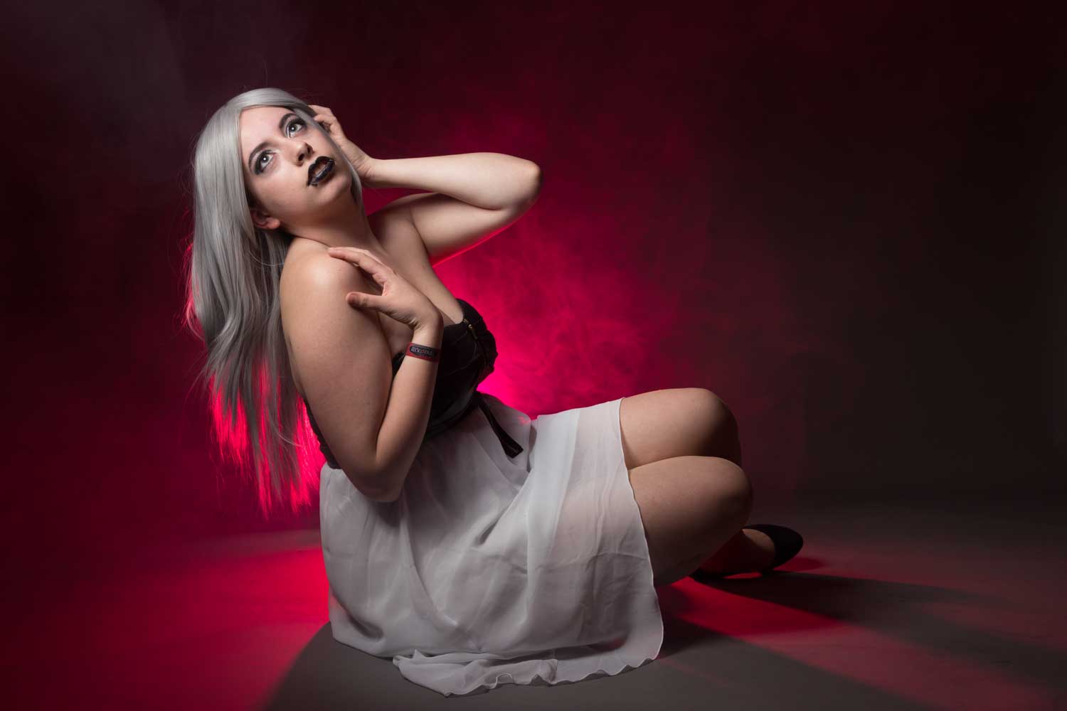 Woman with silver hair and dark makeup poses dramatically seated on floor against a smoky red background.