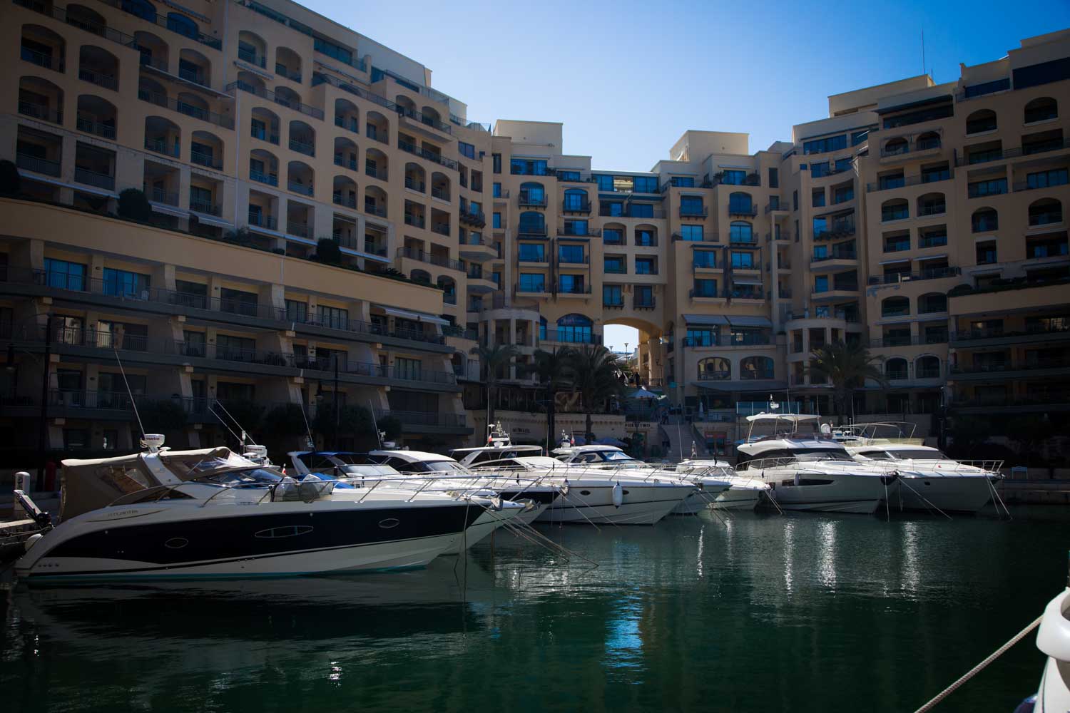 Luxurious yachts docked in a marina, surrounded by modern waterfront buildings under a clear blue sky.