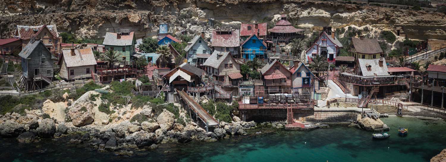 Colorful coastal village with rustic wooden houses on a rocky shore, near turquoise water, set against a cliff backdrop.