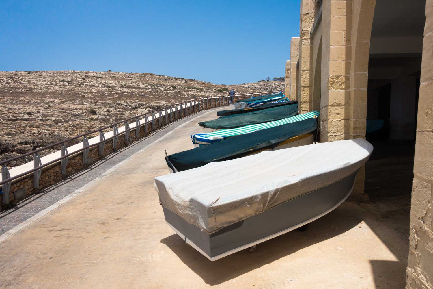Covered boats lined up next to rustic stone buildings on a sunny, coastal path.