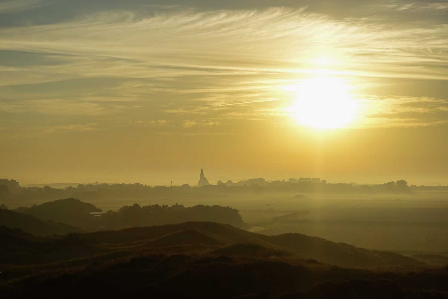 Sun rising over misty fields with a silhouette of a distant village and church spire.