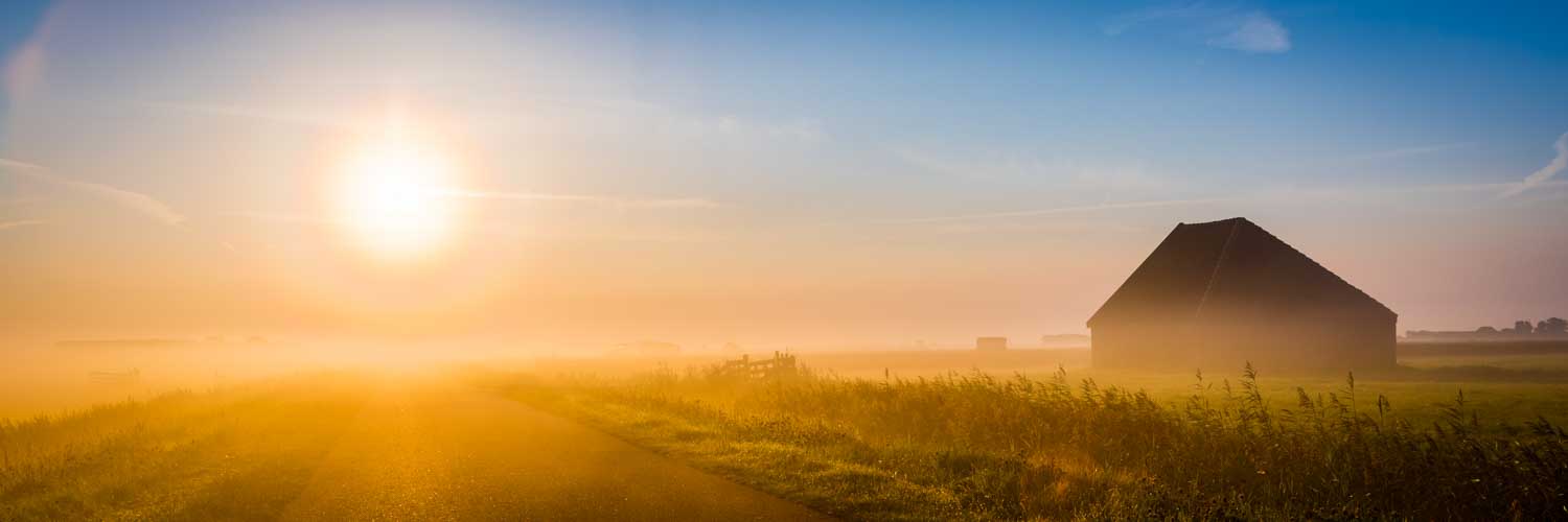 Sunrise over foggy rural landscape with a barn and grassy fields, under a clear blue sky.