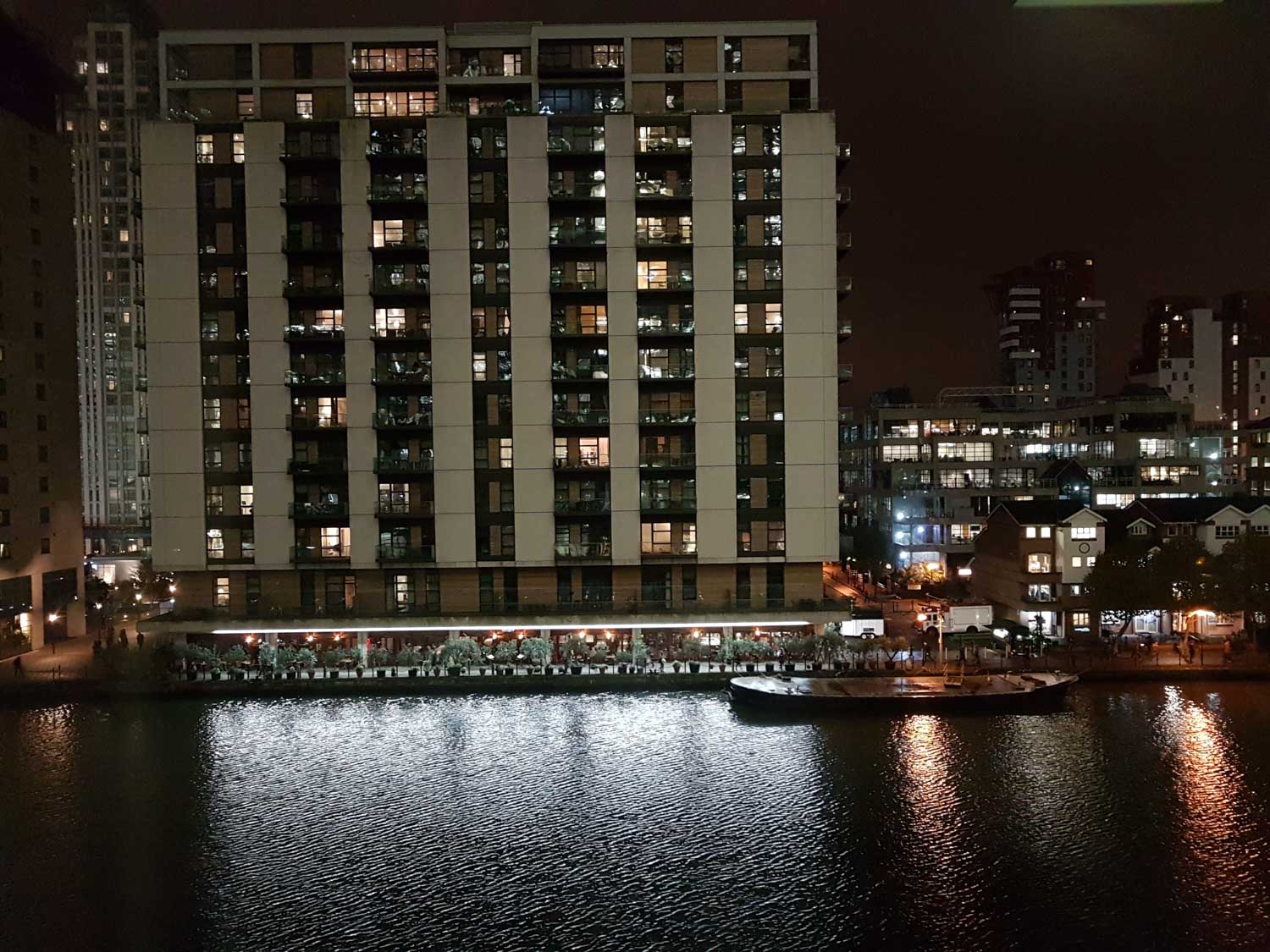 High-rise building with illuminated windows reflecting on a city river at night. Adjacent cityscape and waterfront visible.