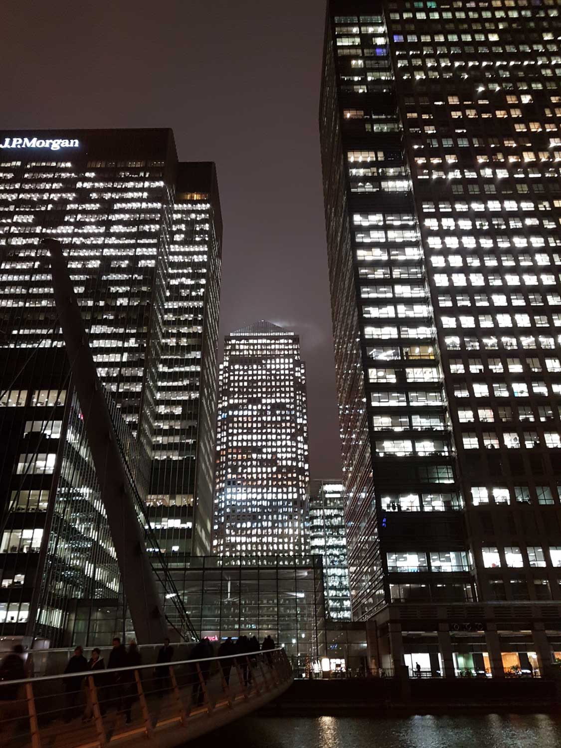 Night view of tall office buildings with bright lights and a pedestrian bridge in an urban setting.