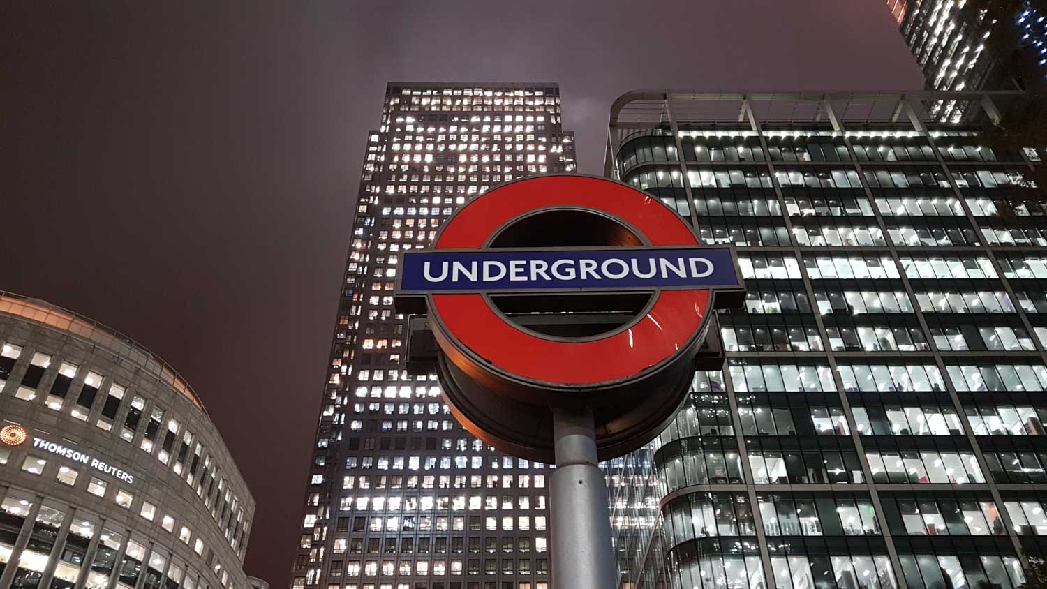 London Underground sign with skyscrapers in the background at night, including a Thomson Reuters building.