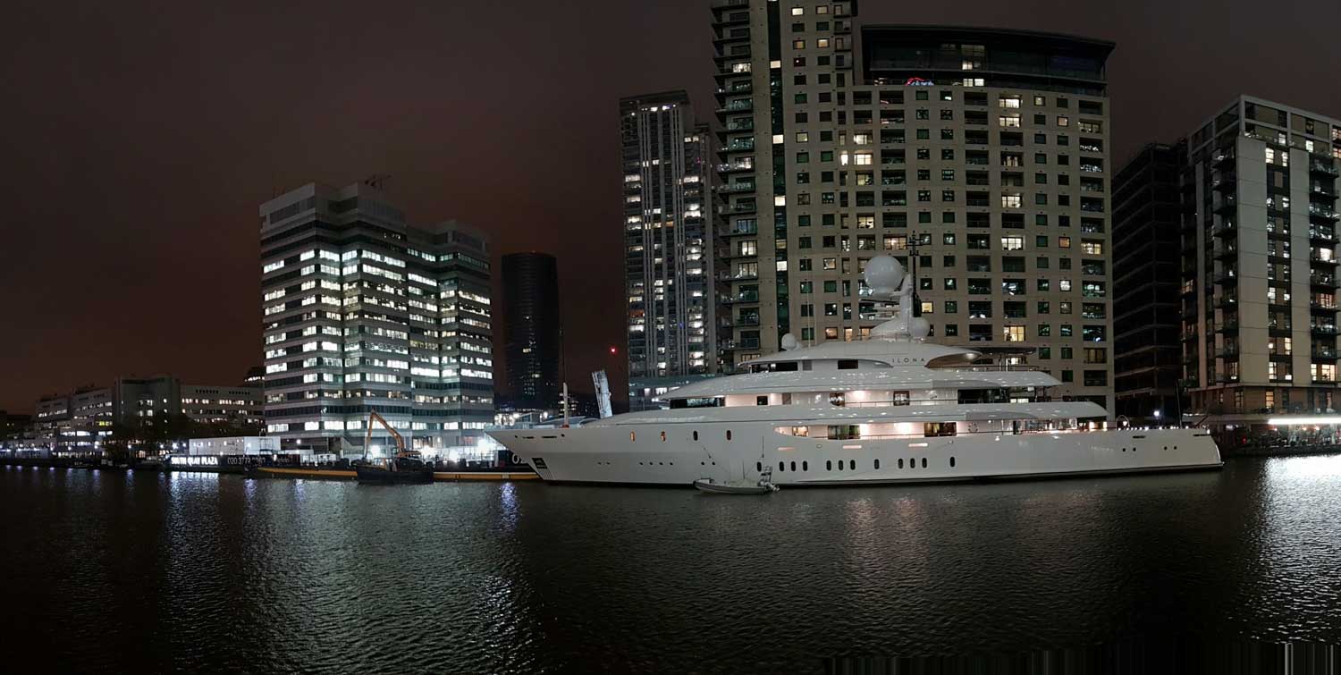 Luxurious white yacht docked at night against a backdrop of illuminated skyscrapers and calm water reflections.