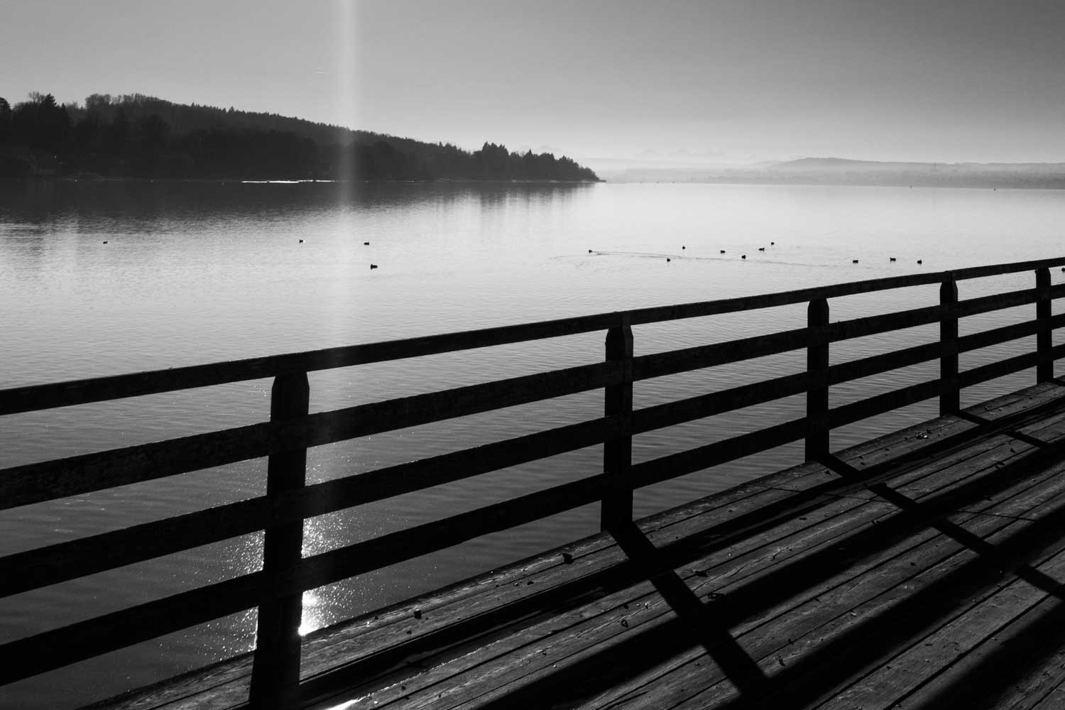 Silhouette of a wooden pier railing overlooking a tranquil lake with gentle hills in the background.