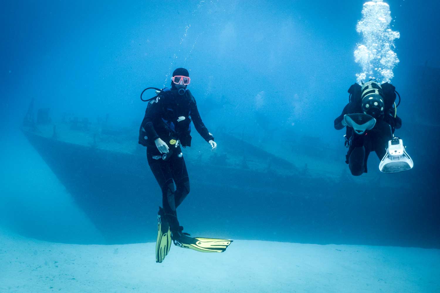 Scuba divers explore a sunken shipwreck in clear blue ocean waters, wearing yellow fins and diving gear.