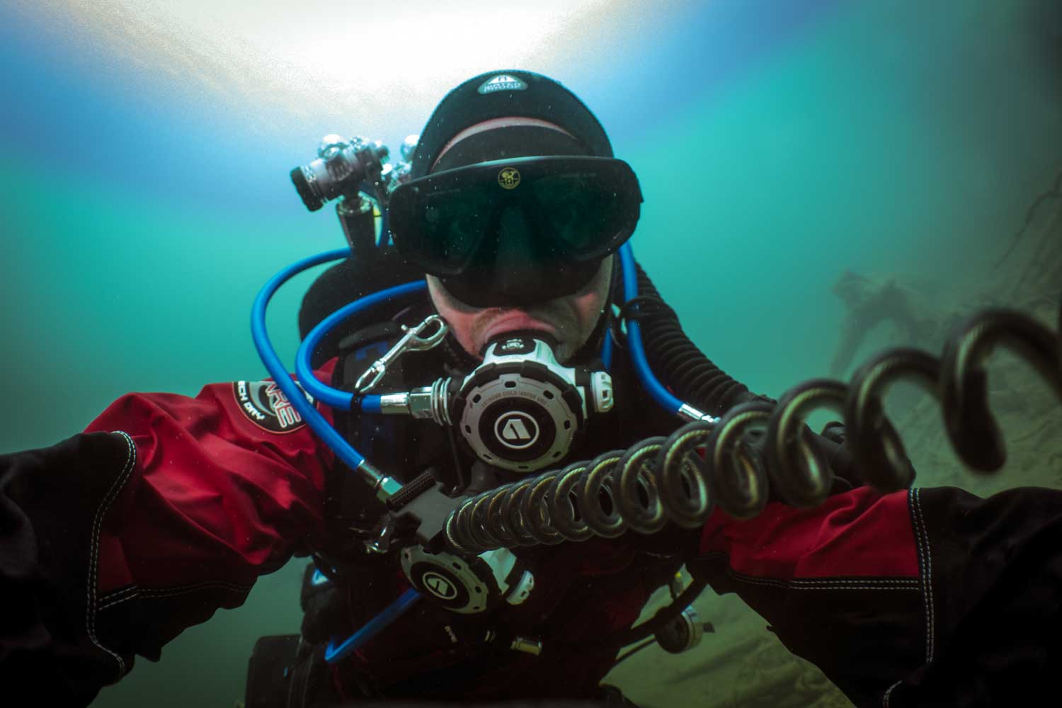 Scuba diver in red drysuit underwater, using a rebreather and capturing a selfie in greenish-blue water.