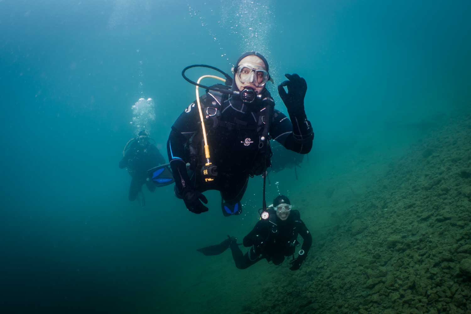Scuba diver underwater giving an OK sign, with two divers in the background near rocky seabed.
