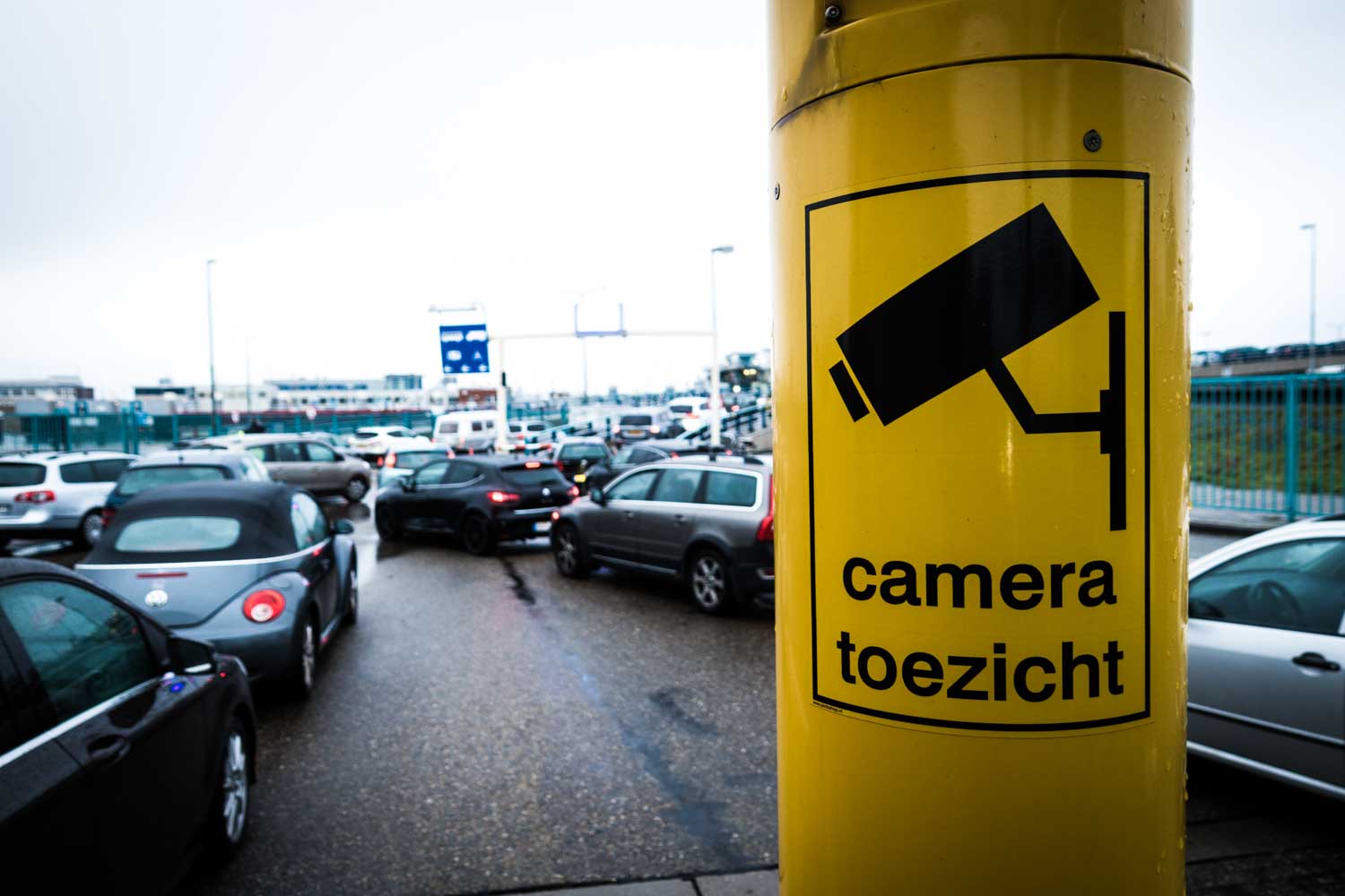Traffic jam at security-monitored area with yellow CCTV camera sign on post, labeled camera toezicht.