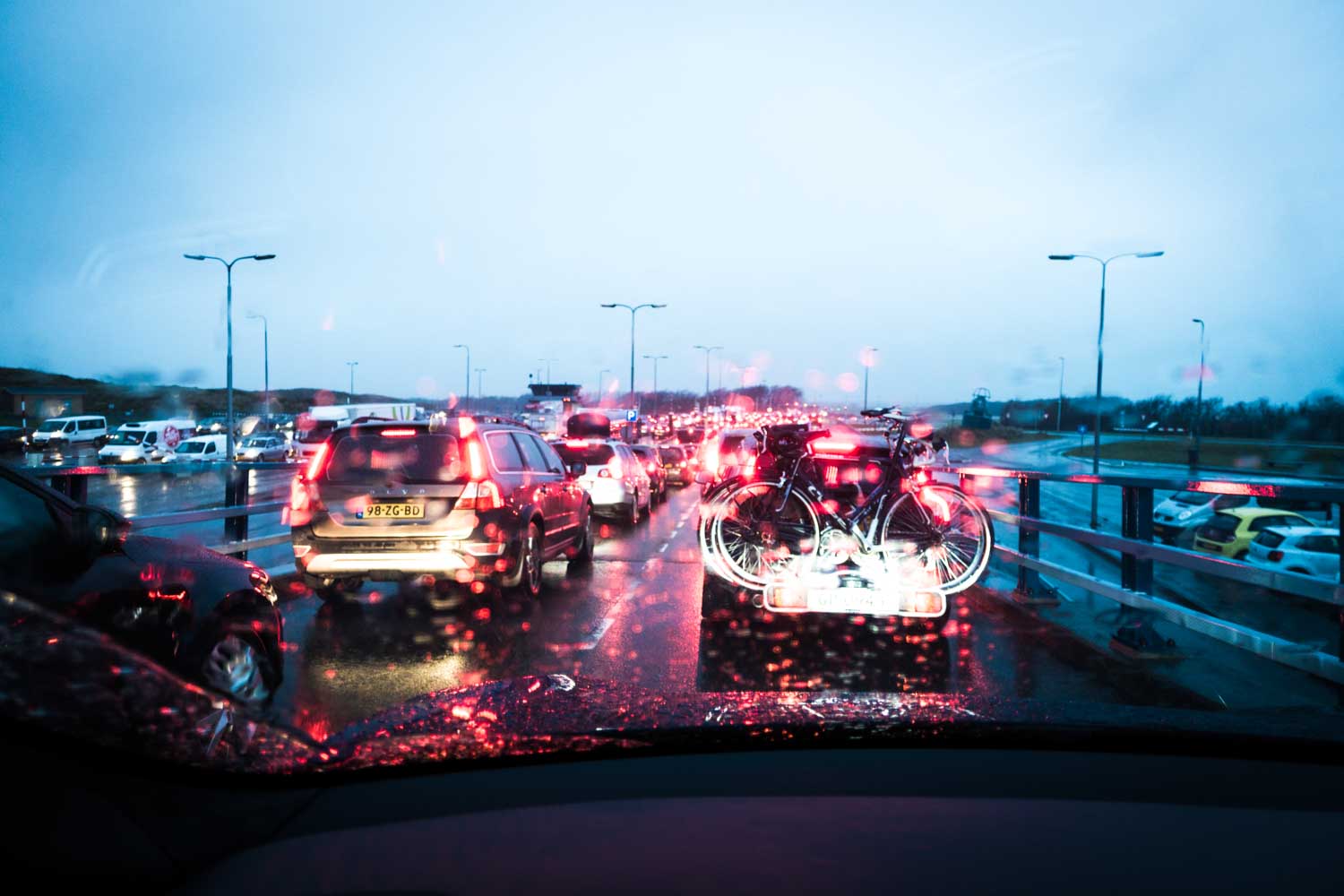 Heavy traffic on a rainy highway at dusk, with cars showing red brake lights and bicycles mounted on a vehicle.