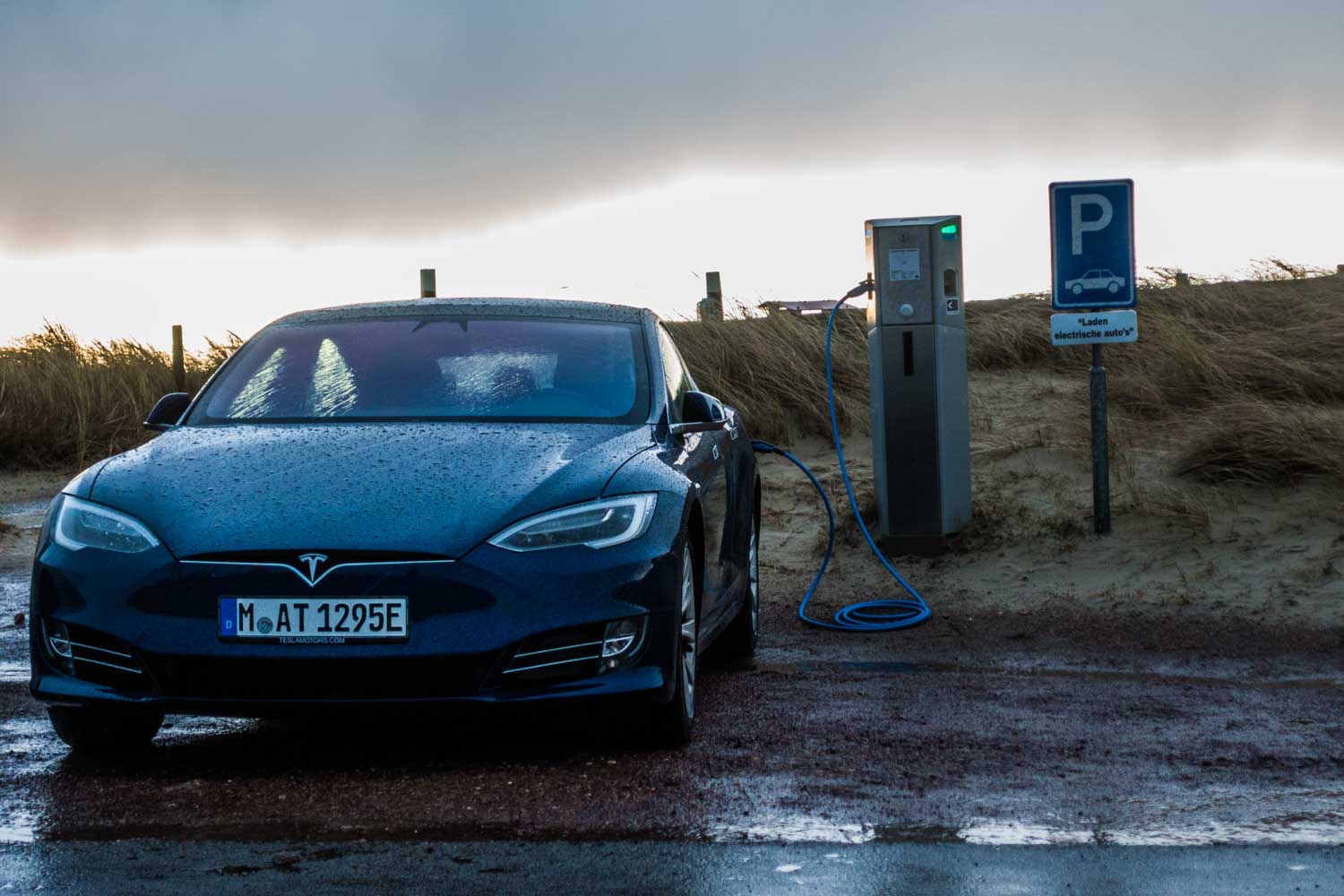Electric car charging at station by sandy roadside under cloudy sky.