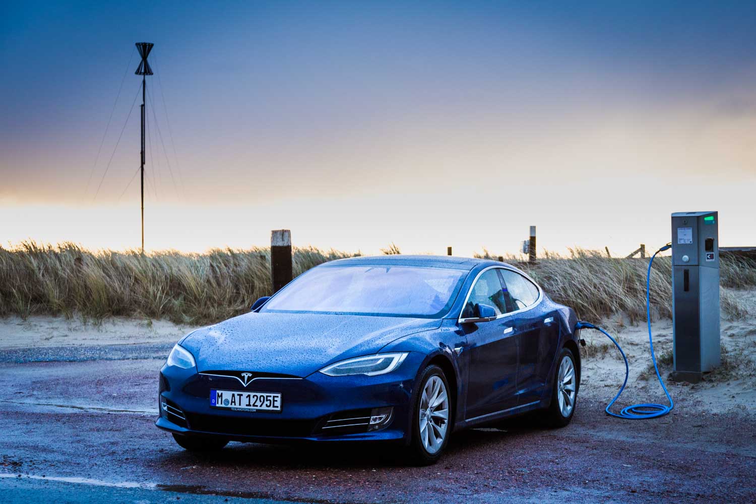 Blue Tesla Model S charging at a beachside station during sunset, with sand dunes and grasses in the background.