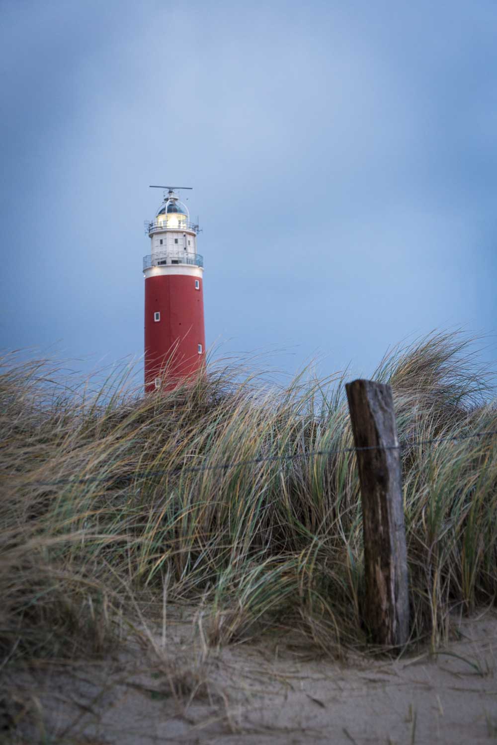 Red lighthouse towering above grassy dunes under a cloudy sky.