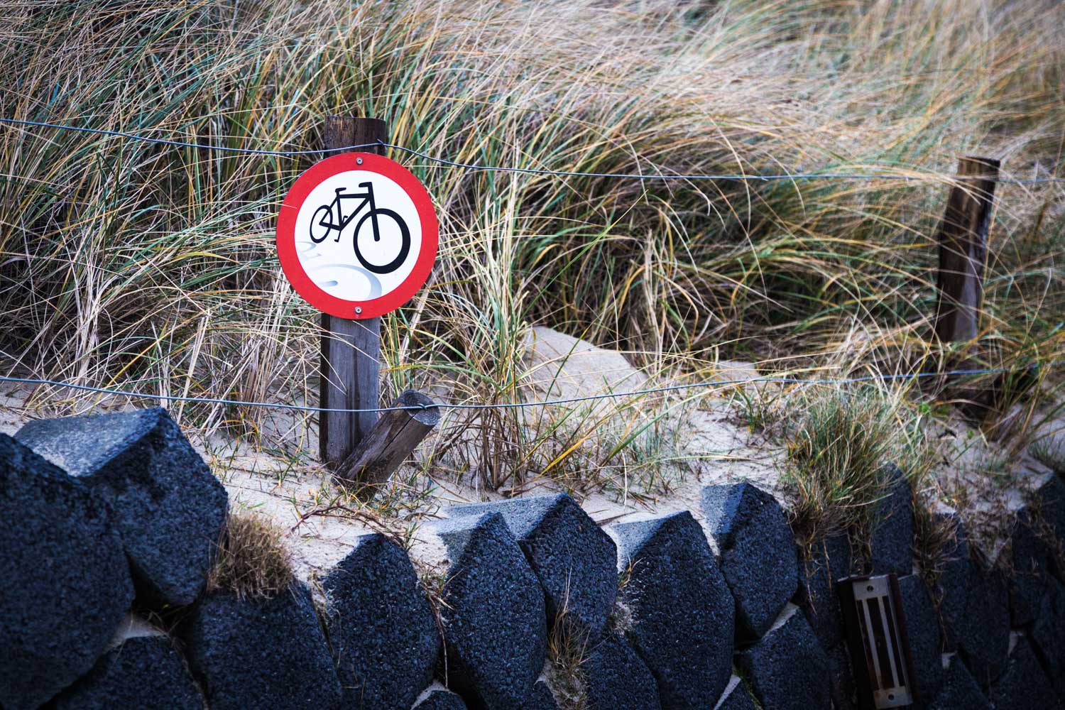 No bicycles sign on sandy path with dune grass and stone barrier in background.