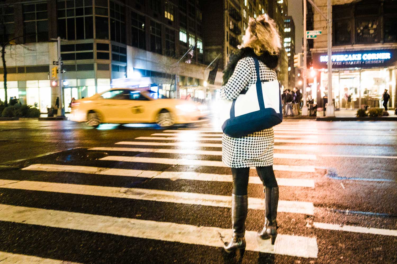 Woman in checkered coat crossing busy city street at night with yellow taxi speeding past.