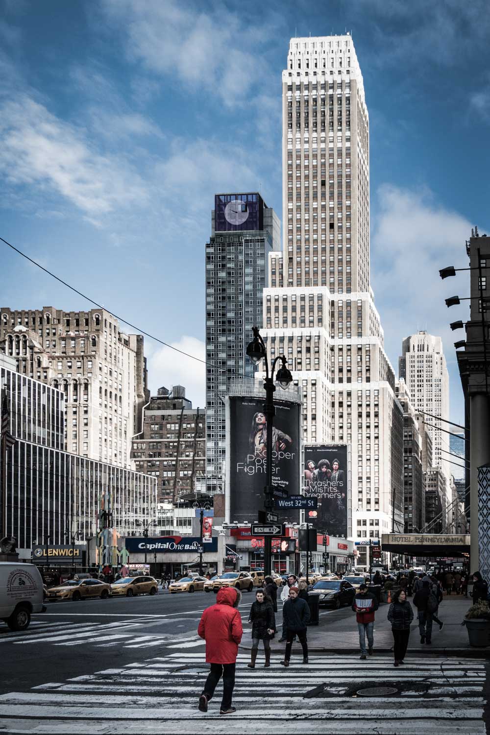 Pedestrians crossing a busy New York City street with tall skyscrapers and taxis under a clear blue sky.