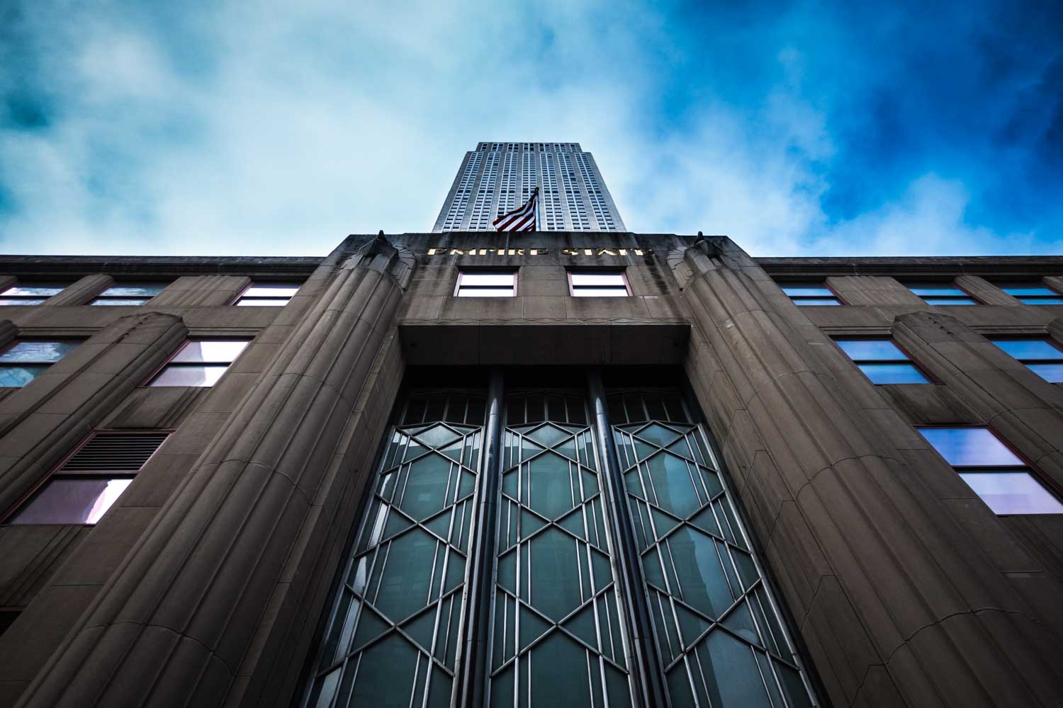 Upward view of the Empire State Building entrance against a cloudy blue sky.