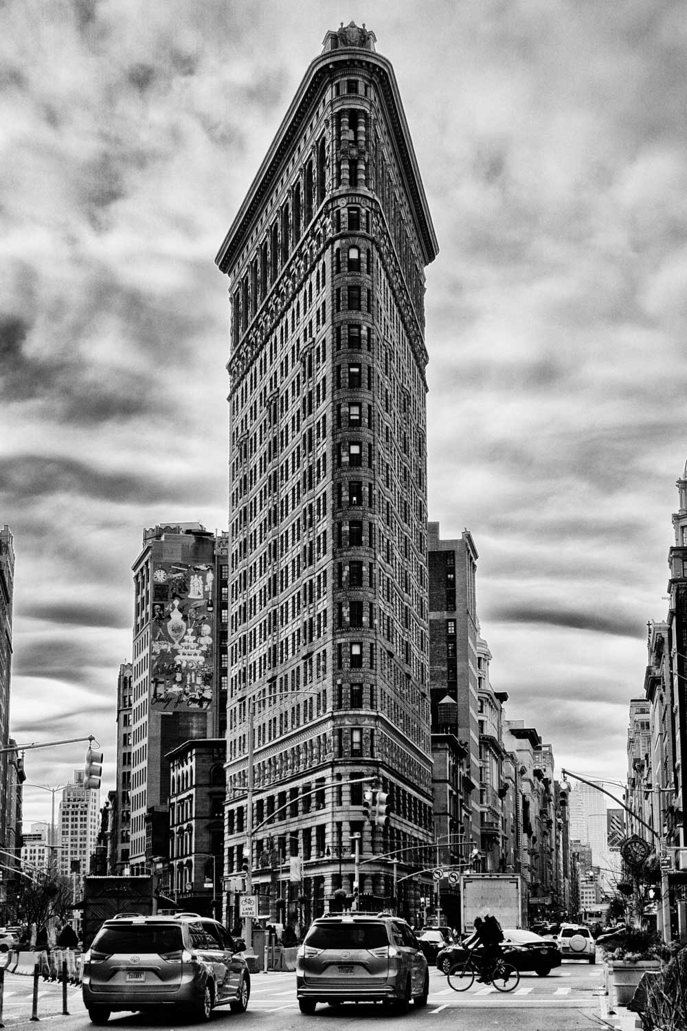 Black and white photo of the Flatiron Building in New York City with cars and cyclists on the street below.