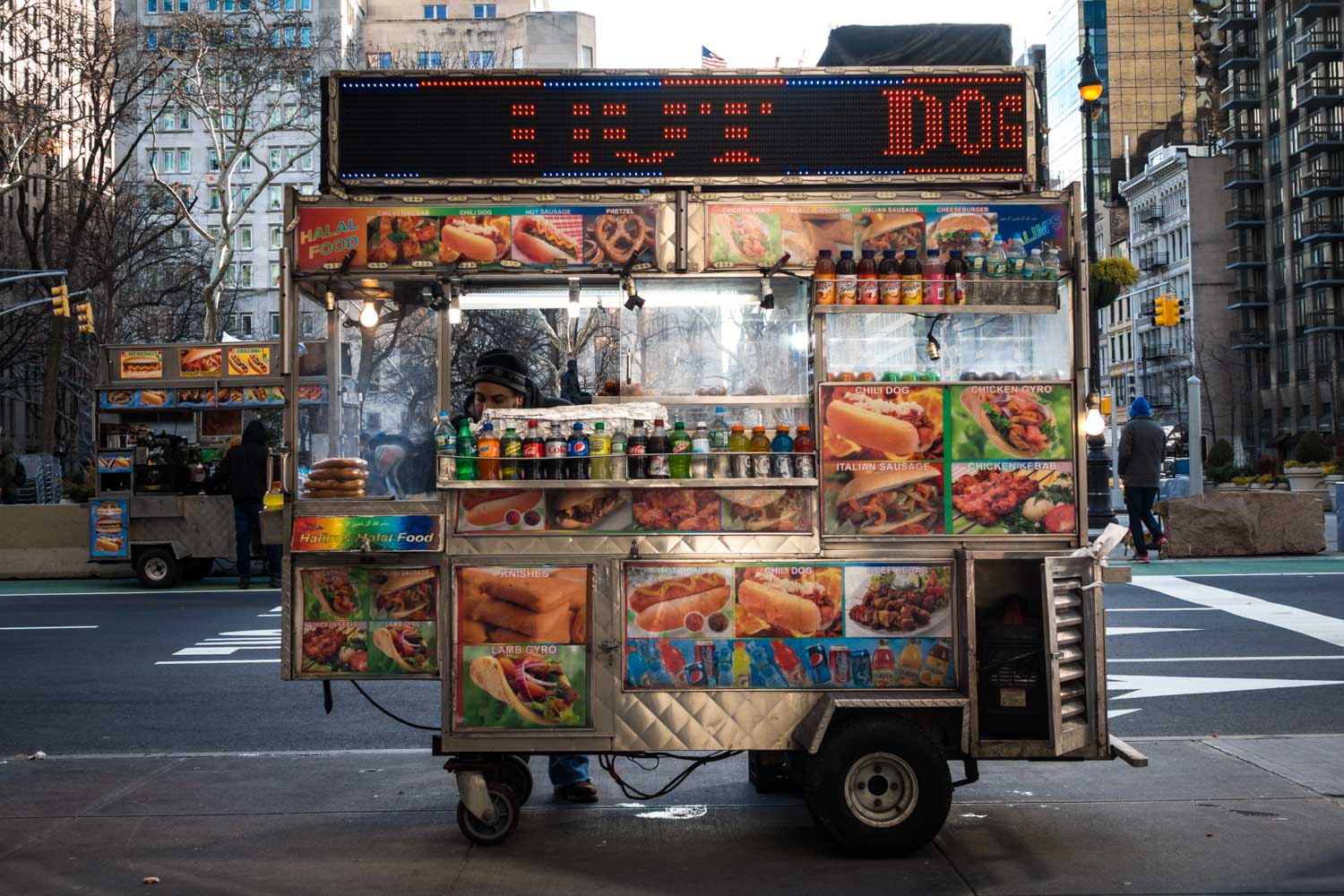 Street food cart with colorful menu of hot dogs, gyros, and drinks set against an urban city backdrop.