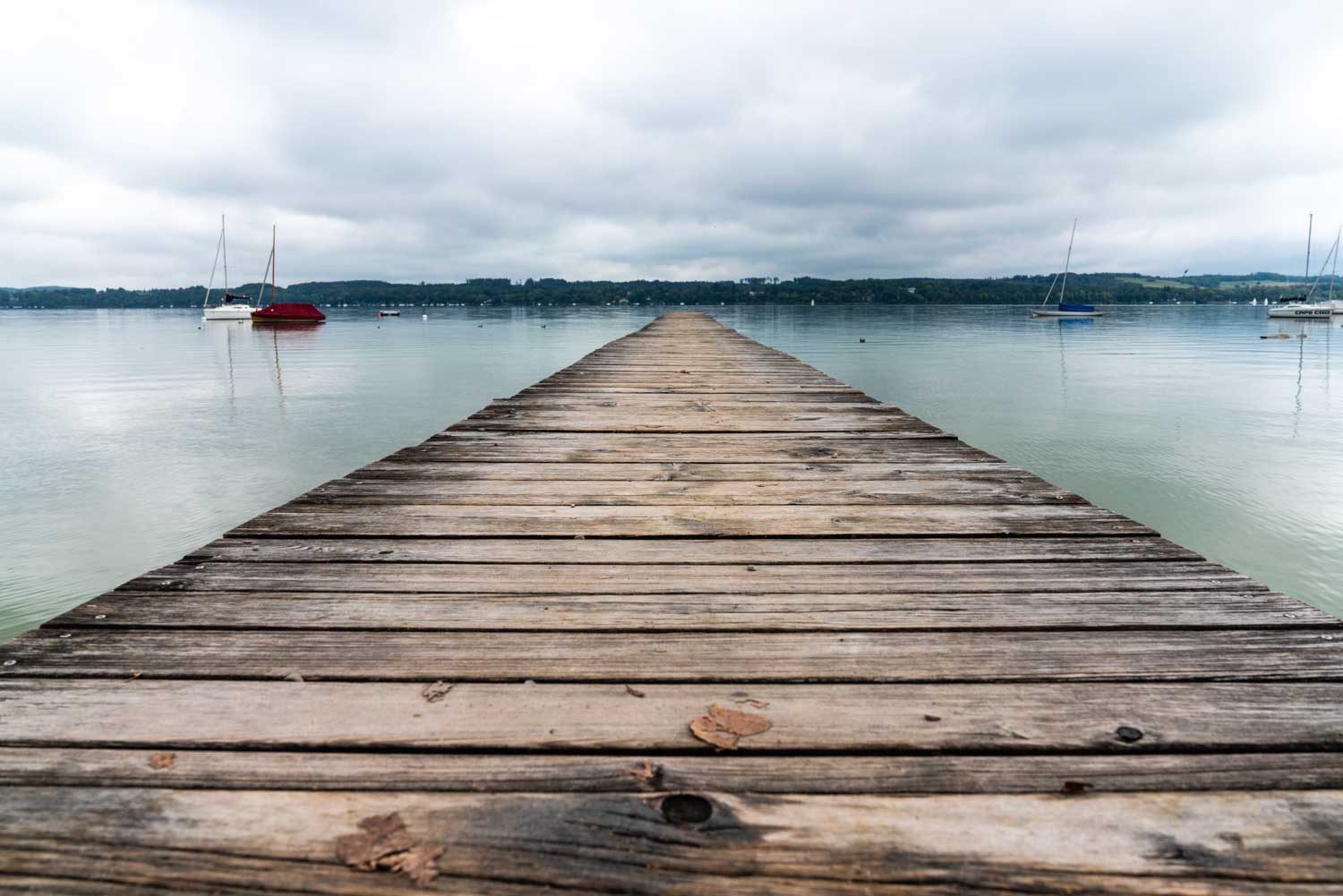 Wooden pier extending over a calm lake with sailboats and cloudy sky in the background.