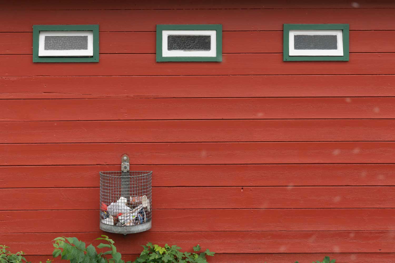 Red wooden wall with three small windows, green frames, and an attached metal trash bin filled with garbage.