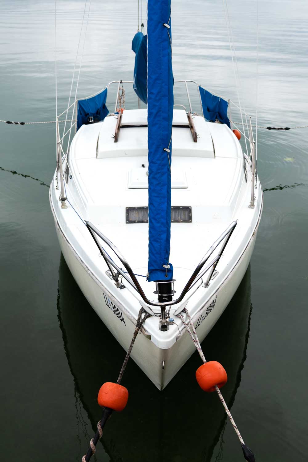 Front view of a docked sailboat with blue sail cover and orange buoys on calm water.
