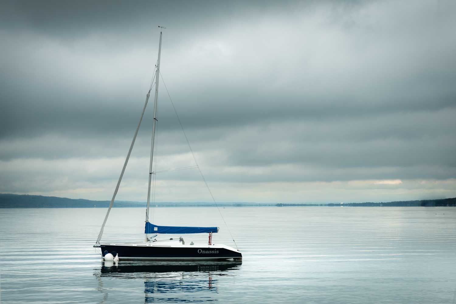Sailboat with blue cover floating on calm lake under cloudy skies.