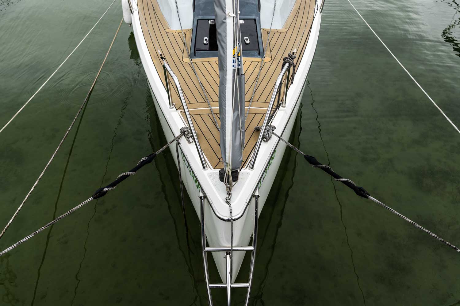 Bow of a docked sailboat with wooden decking, secured with ropes on calm green water.