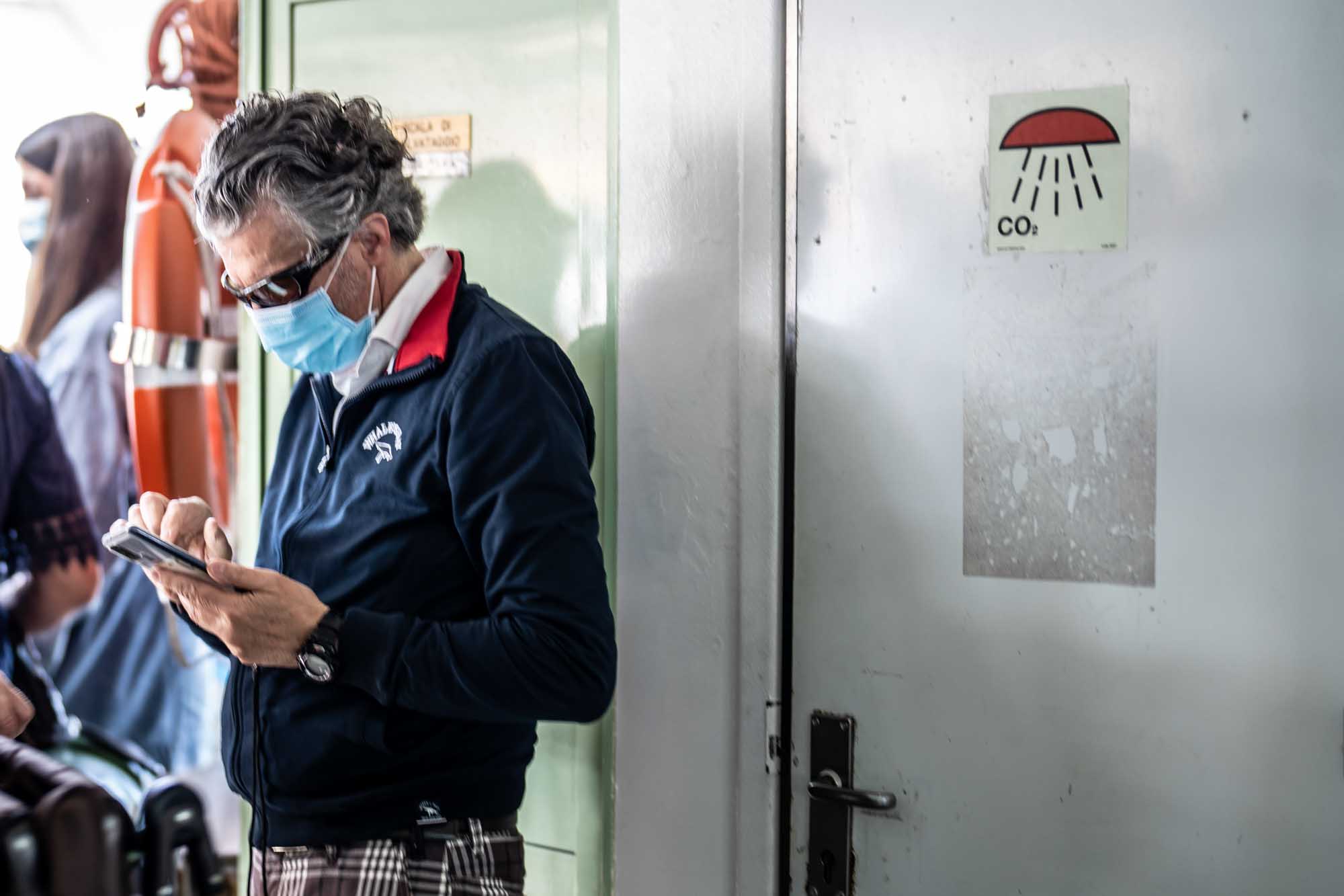 Man wearing mask and sunglasses uses phone indoors near CO2 sign and life jacket on wall.