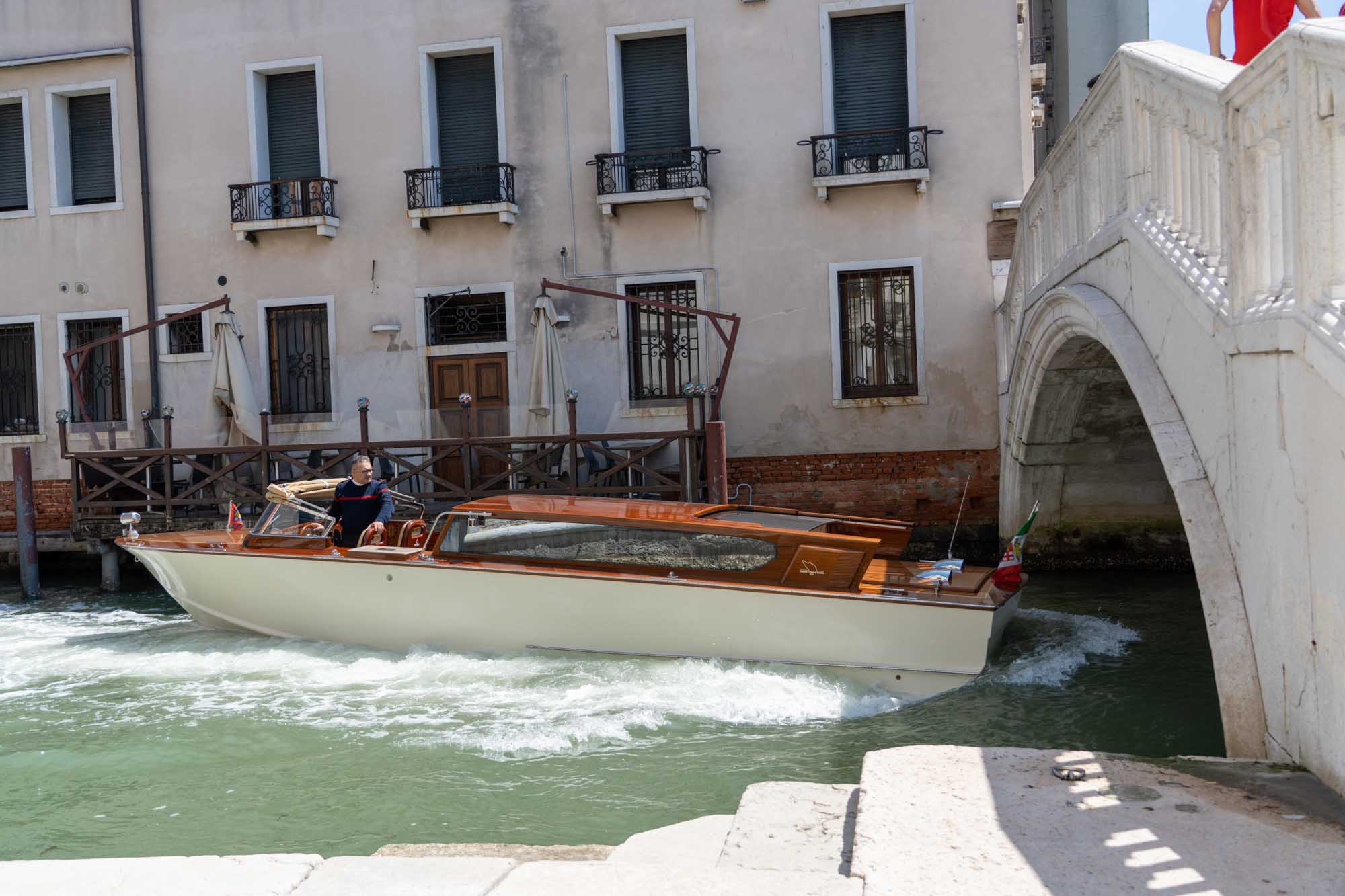 A motorboat navigates a Venetian canal under a stone bridge, near a historic building with shuttered windows.