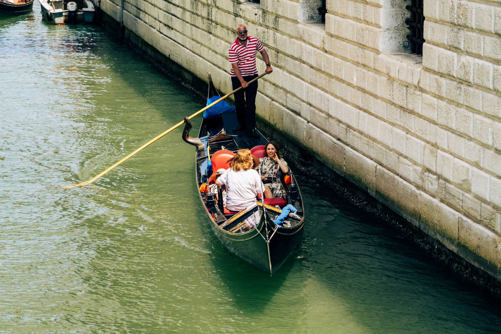 Gondolier rowing tourists in Venetian canal beside stone building under bright sunlight.