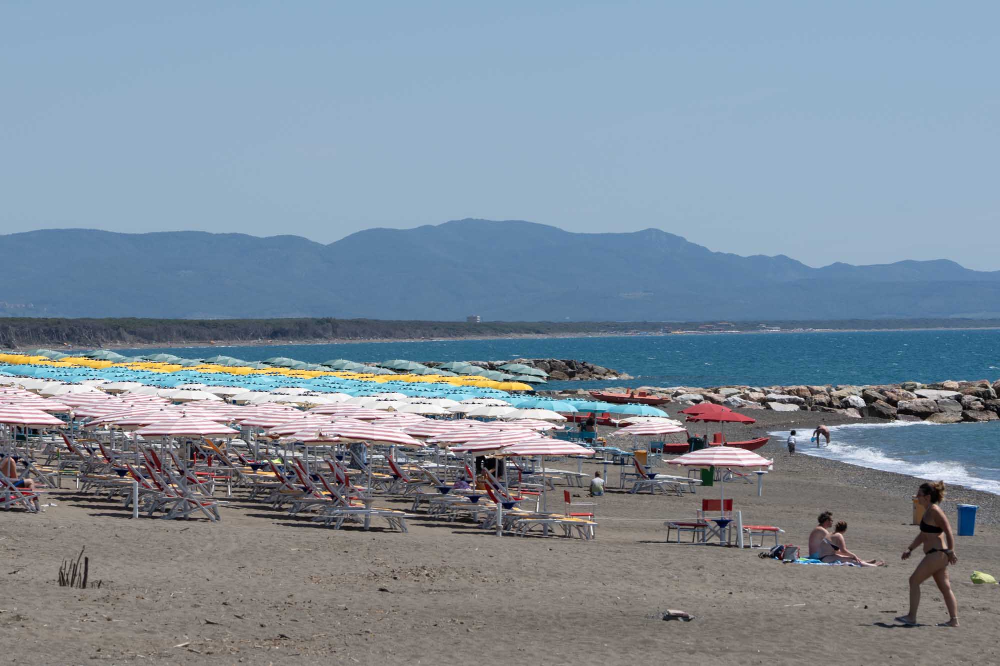 Sunny beach with colorful umbrellas and deck chairs, bordered by a mountainous horizon and blue sea waves.