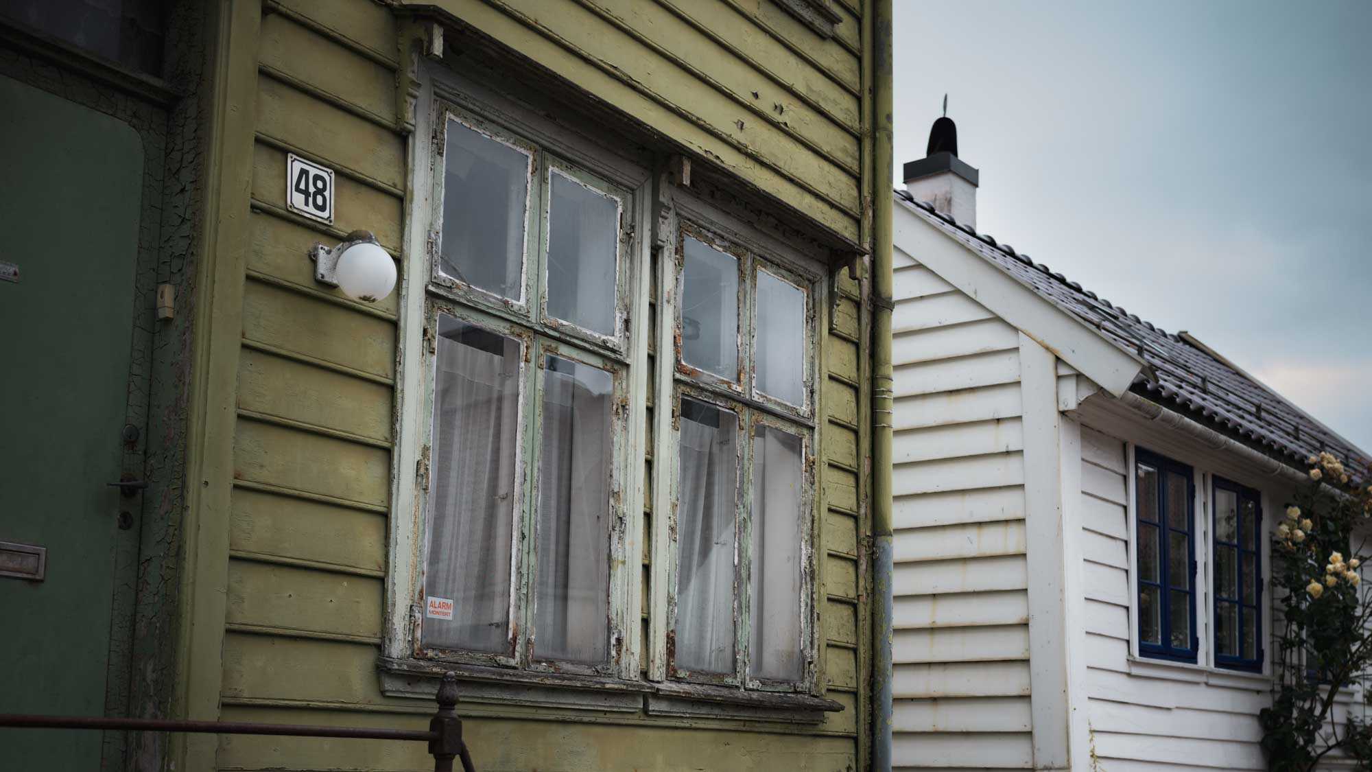 Old green wooden house with peeling paint and number 48 beside a white house with blue windows and flowers.