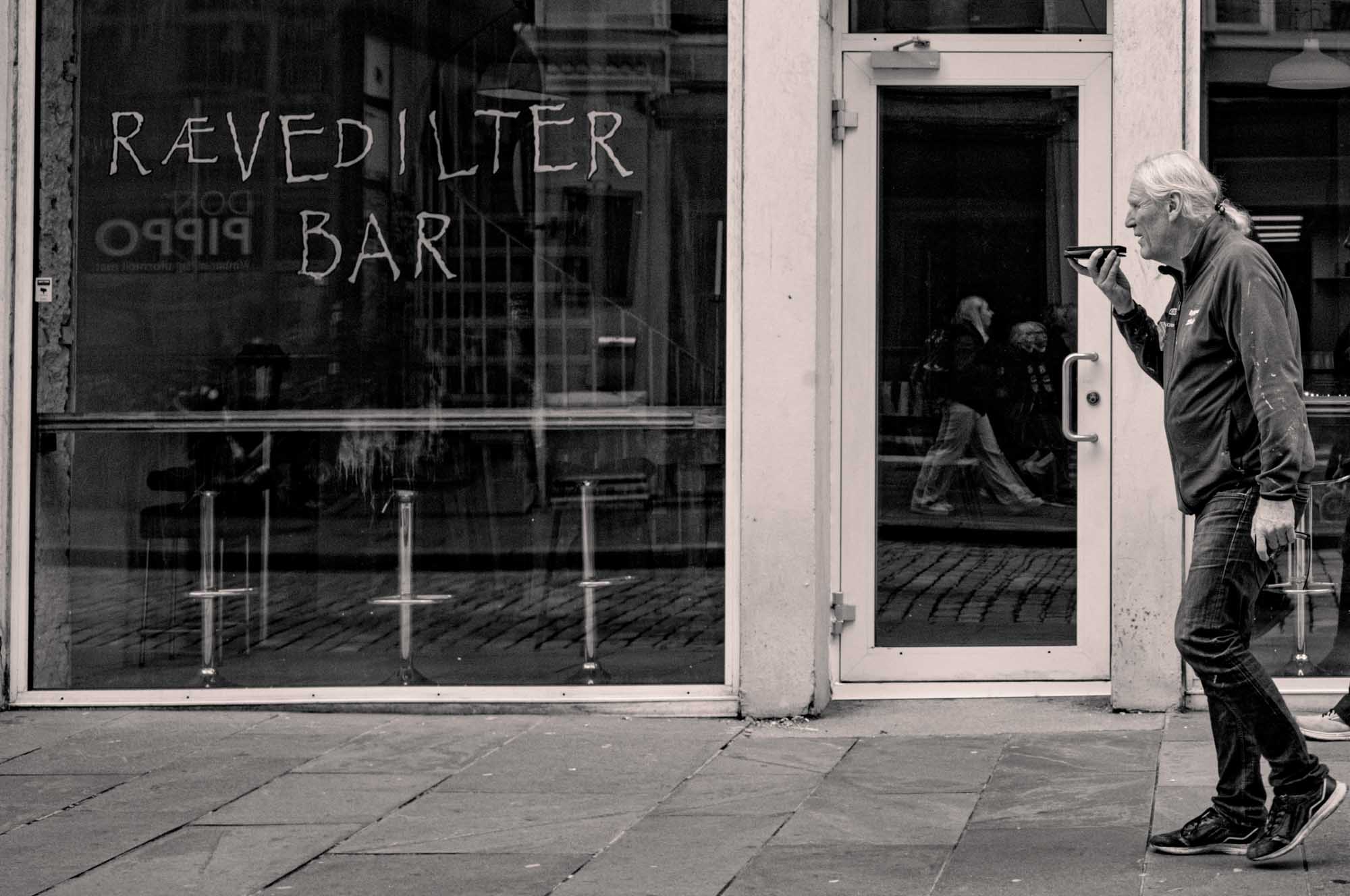 Man using smartphone while walking past a bar with handwritten sign, reflecting street scene in window, monochrome image.