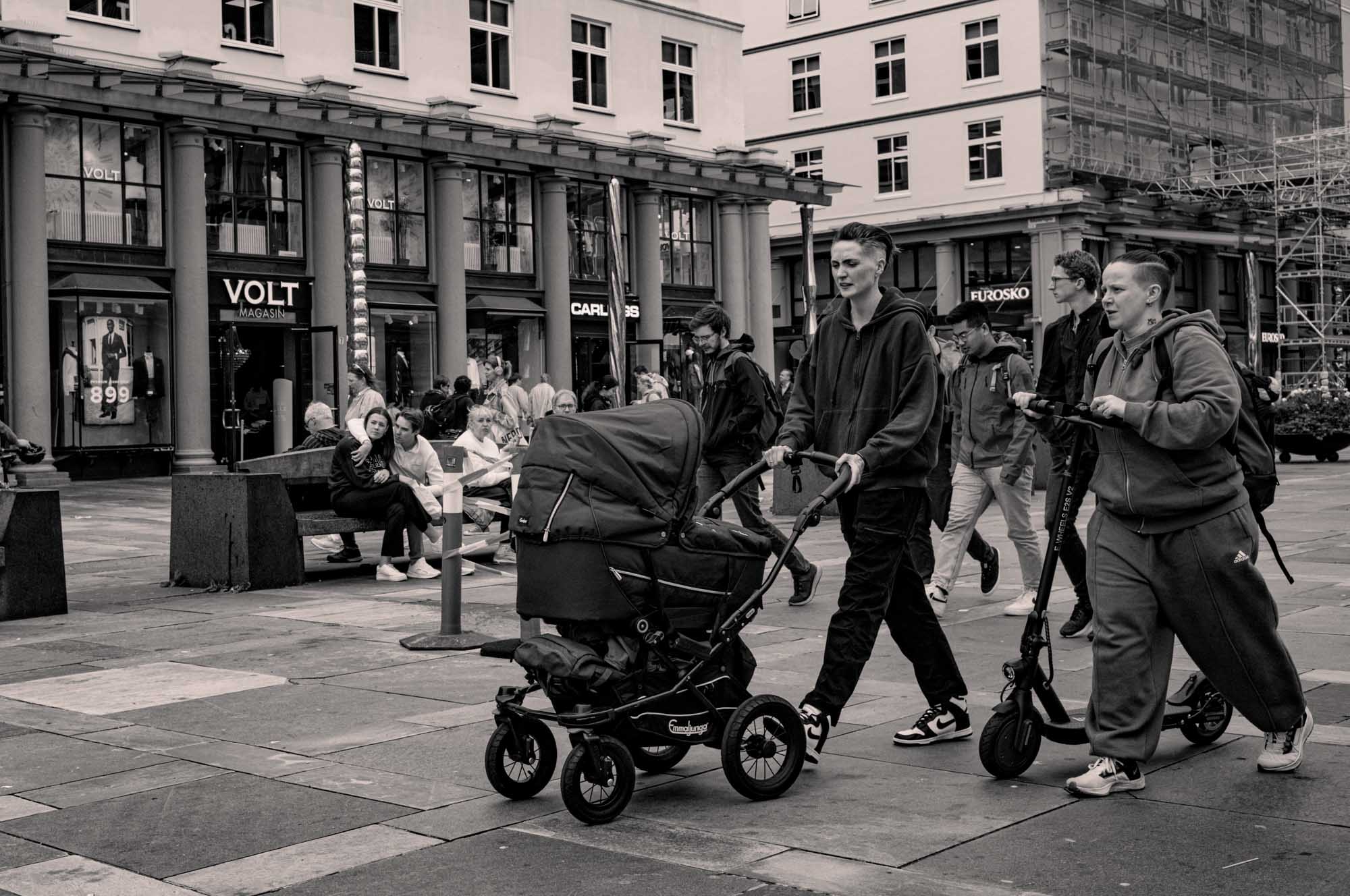 People walking on a city street; one person pushes a stroller, another rides a scooter. Shops and pedestrians in the background.
