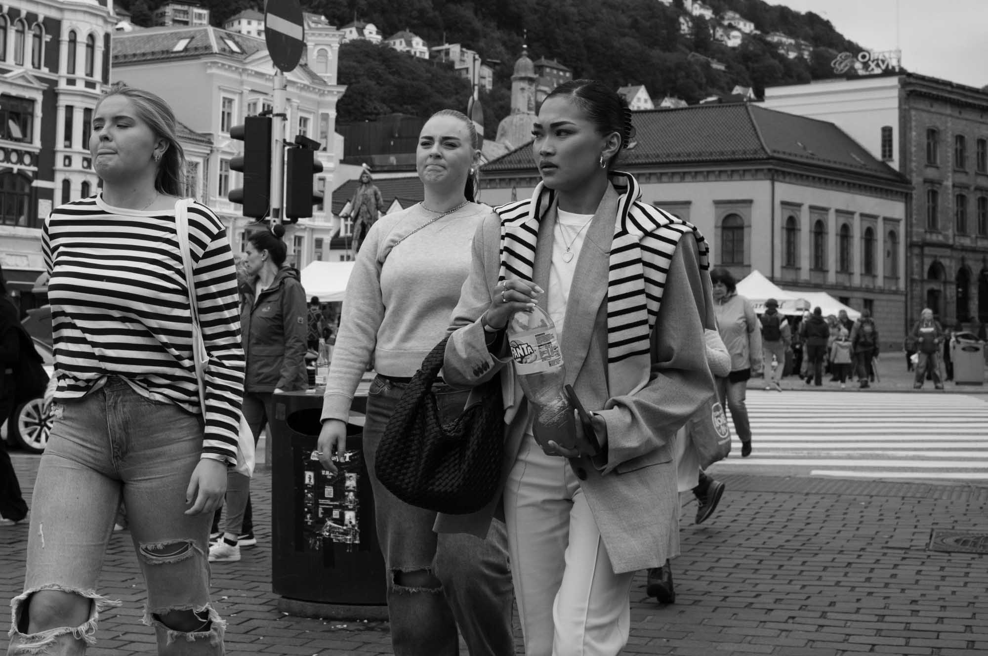 Three women walking in a lively urban street scene, surrounded by historic buildings and people.