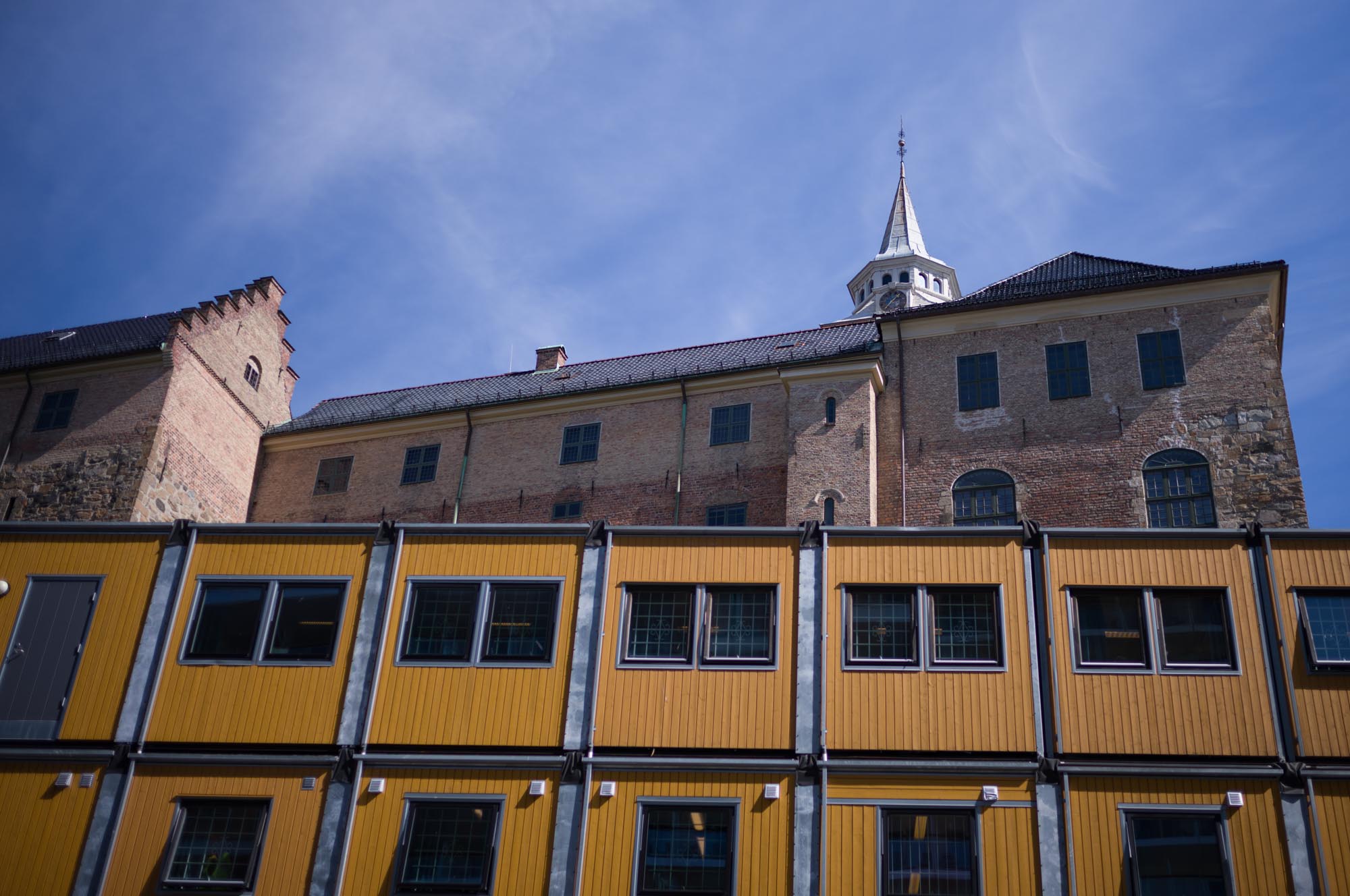 Historic brick building with a spire above modern yellow modular units under a clear blue sky.